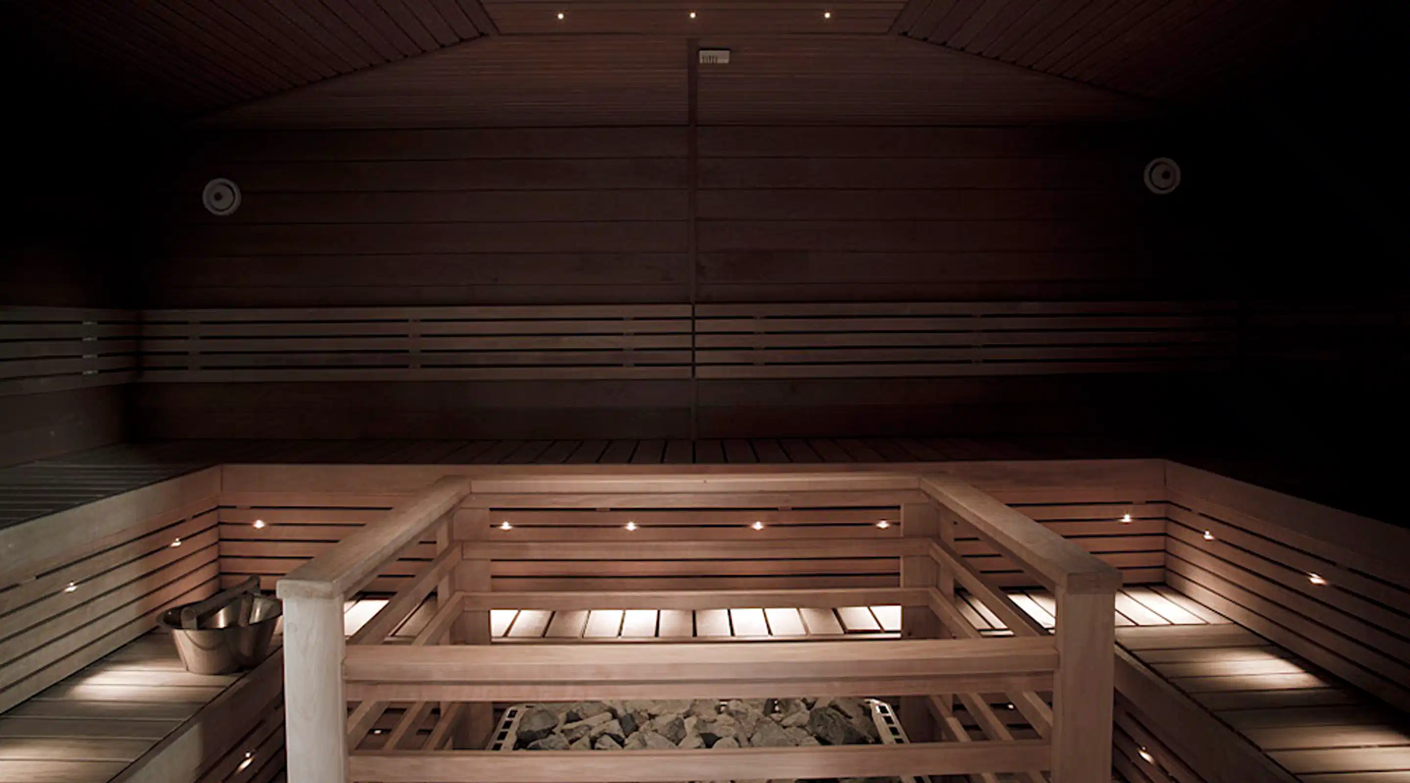 Dimly lit wooden sauna interior. Wooden benches and a heater with sauna stones visible in foreground. Minimalist lighting creates atmosphere.