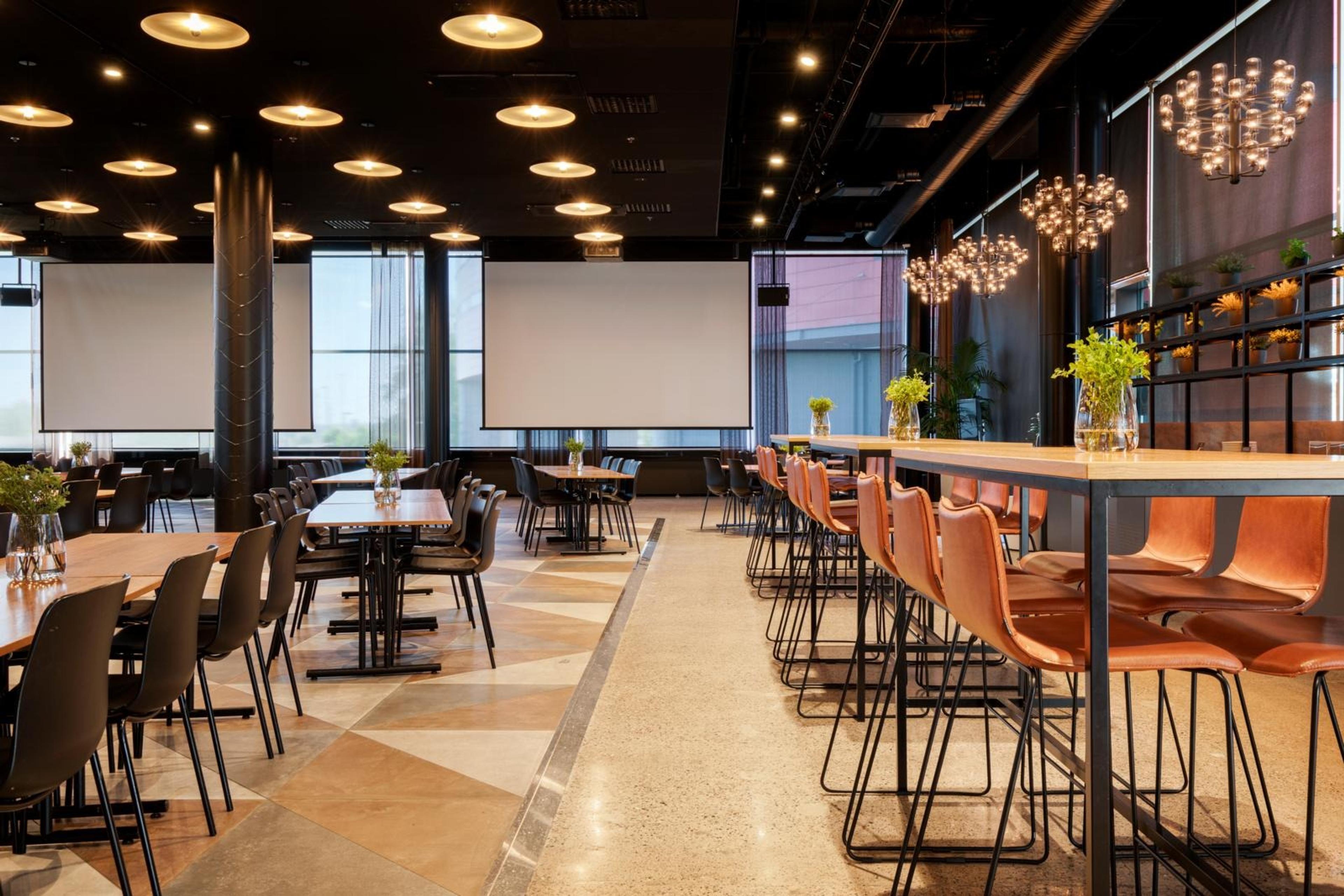 Modern konferens- och evenemangsrestaurang med mörkt tak, ljust golv, vita projektionsdukar och träbord med svarta och orange stolar.
