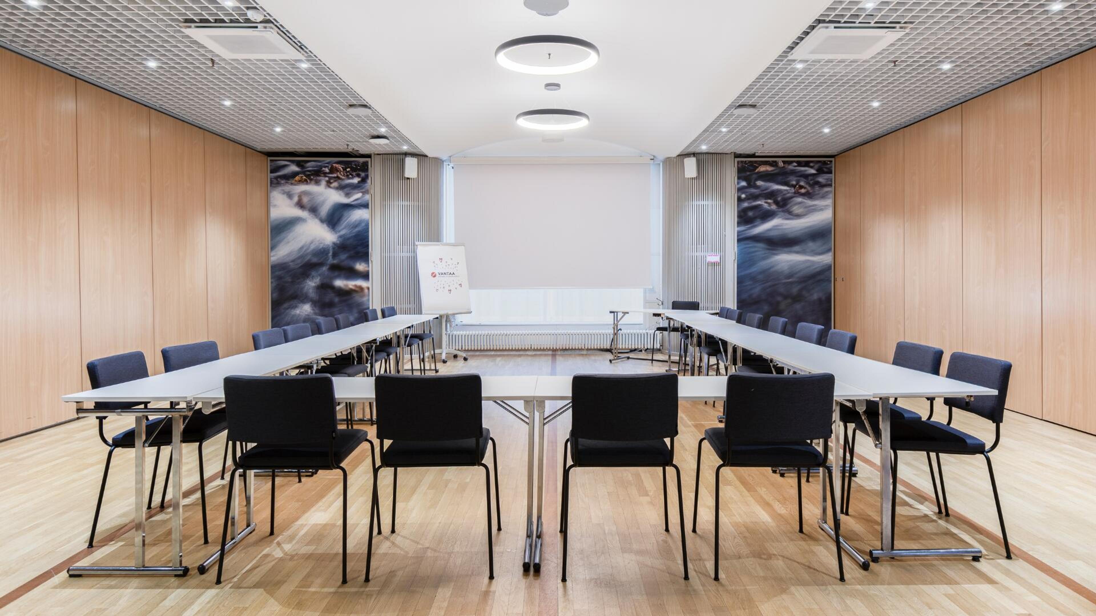 Moderni kokoushuone U-muotoisella pöydällä, mustilla tuoleilla, puisella lattialla ja vesiaiheisilla seinäkuvilla.