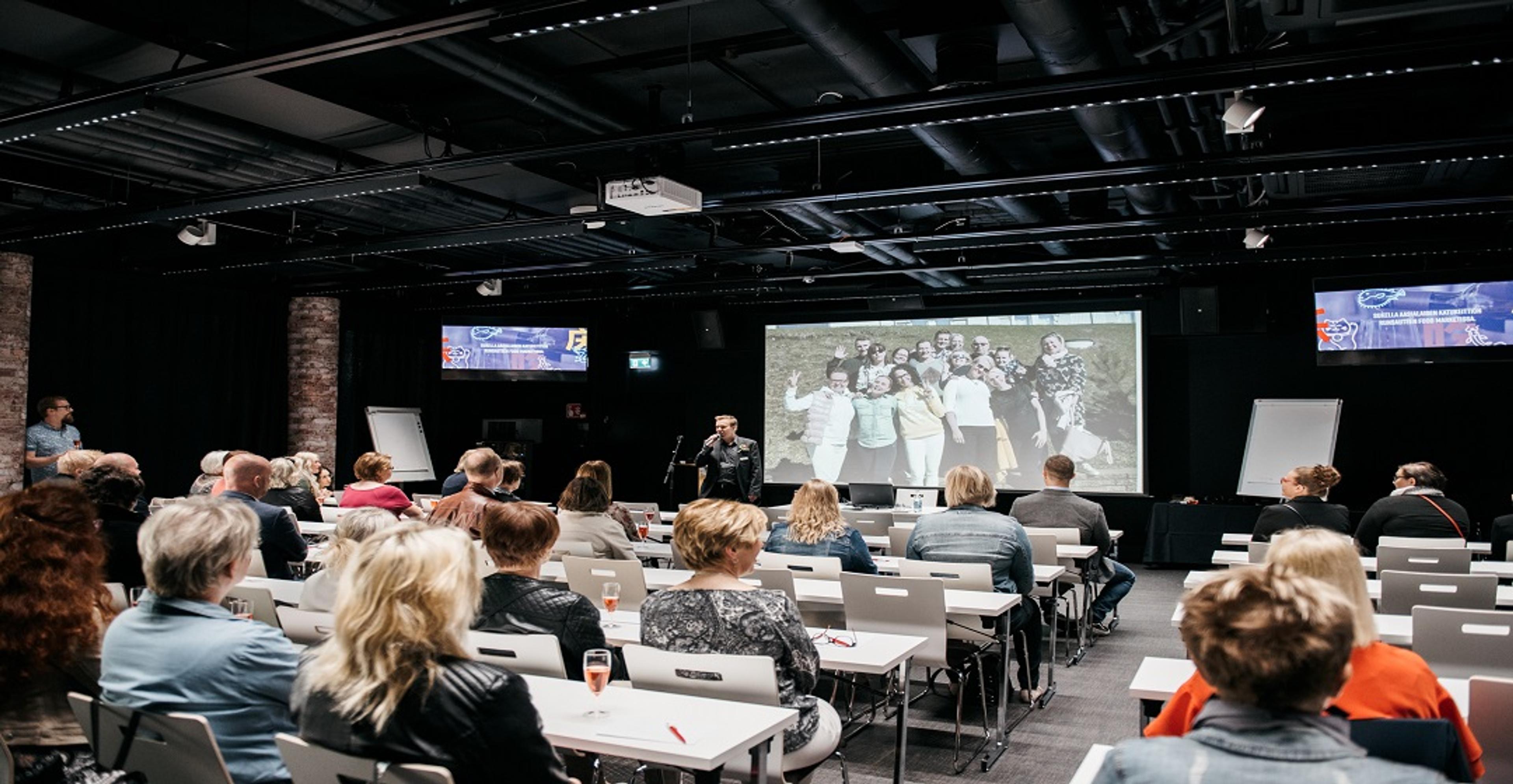 Konferenssihuone, jossa osallistujat istuvat pöytien ääressä kuuntelemassa puhujaa. Seinällä on suuri projektori ja näyttöjä.