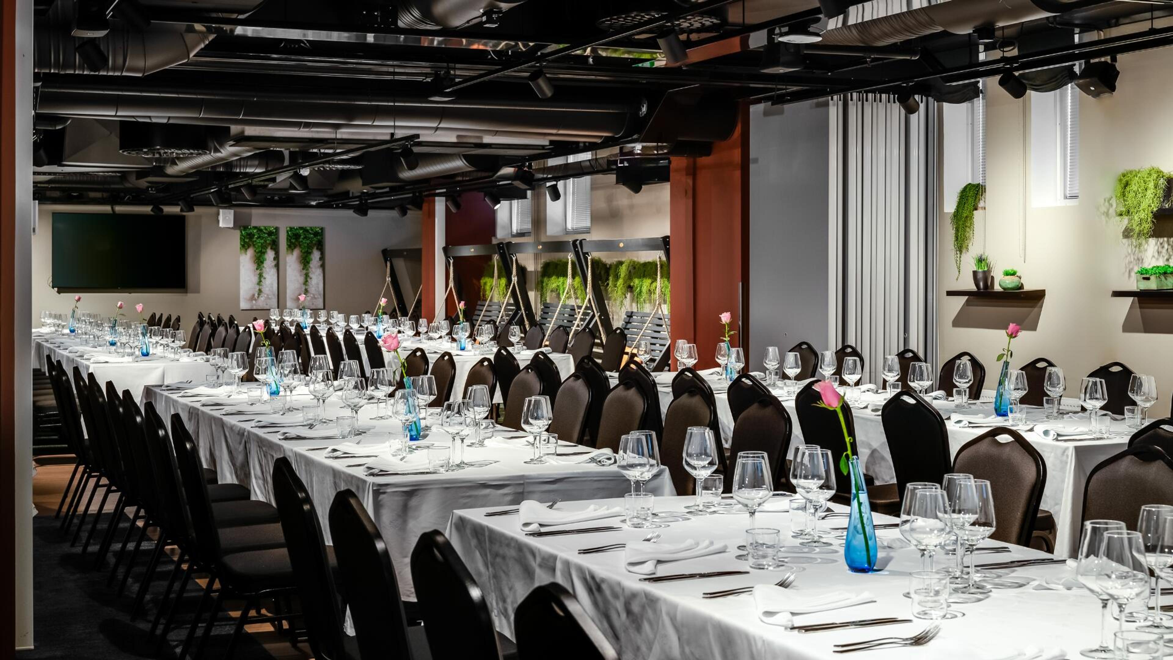 Juhlatila pitkillä pöydillä, jotka on katettu valkoisilla liinoilla, viinilaseilla ja ruokailuvälineillä. Tilassa on viherkasveja ja vaaleanpunaisia kukkia maljakoissa.