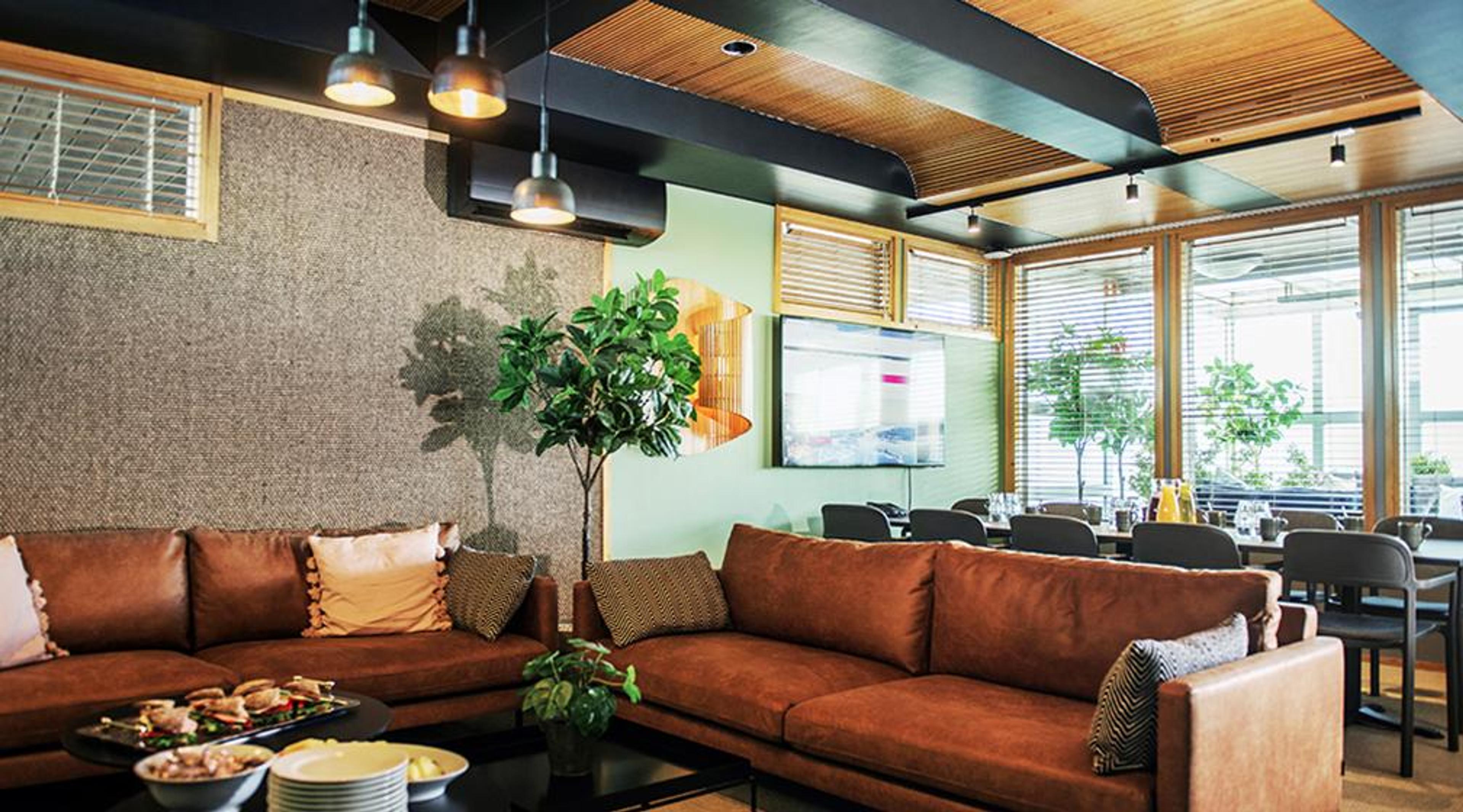 Moderni lounge-tila, jossa ruskea kulmasohva, vihreät kasvit, puukatto ja suuret ikkunat. Taustalla on pöytiä ja tuoleja.