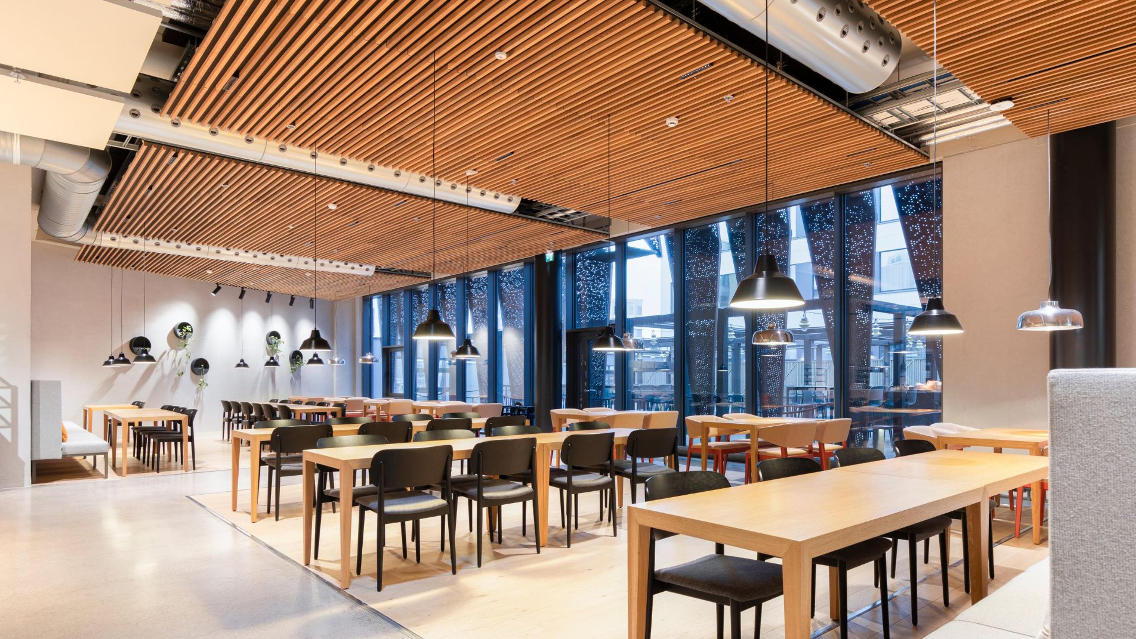 Moderni ravintola puulattiakatolla, valoisilla ikkunoilla ja useilla puupöydillä mustilla tuoleilla. Sileä lattia ja riippuvalaisimet.