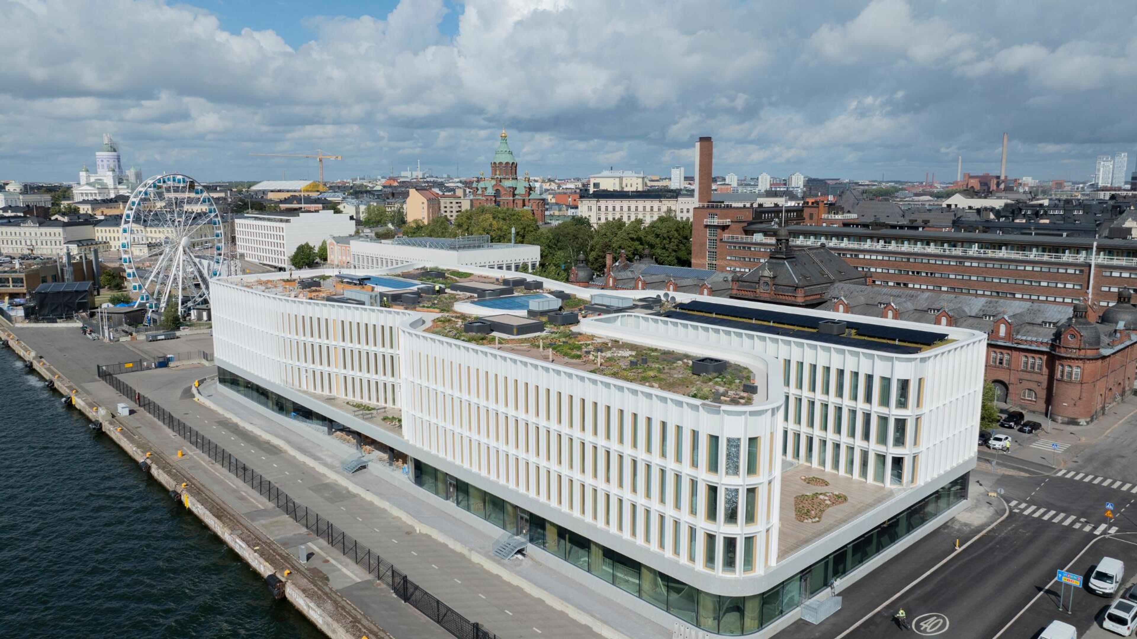 Ilmakuva Katajanokan Laituri rakennuksesta satama-alueella, jolla on viherkatto. Taustalla näkyy Helsingin kaupunkimaisema, maailmanpyörä ja Tuomiokirkko.