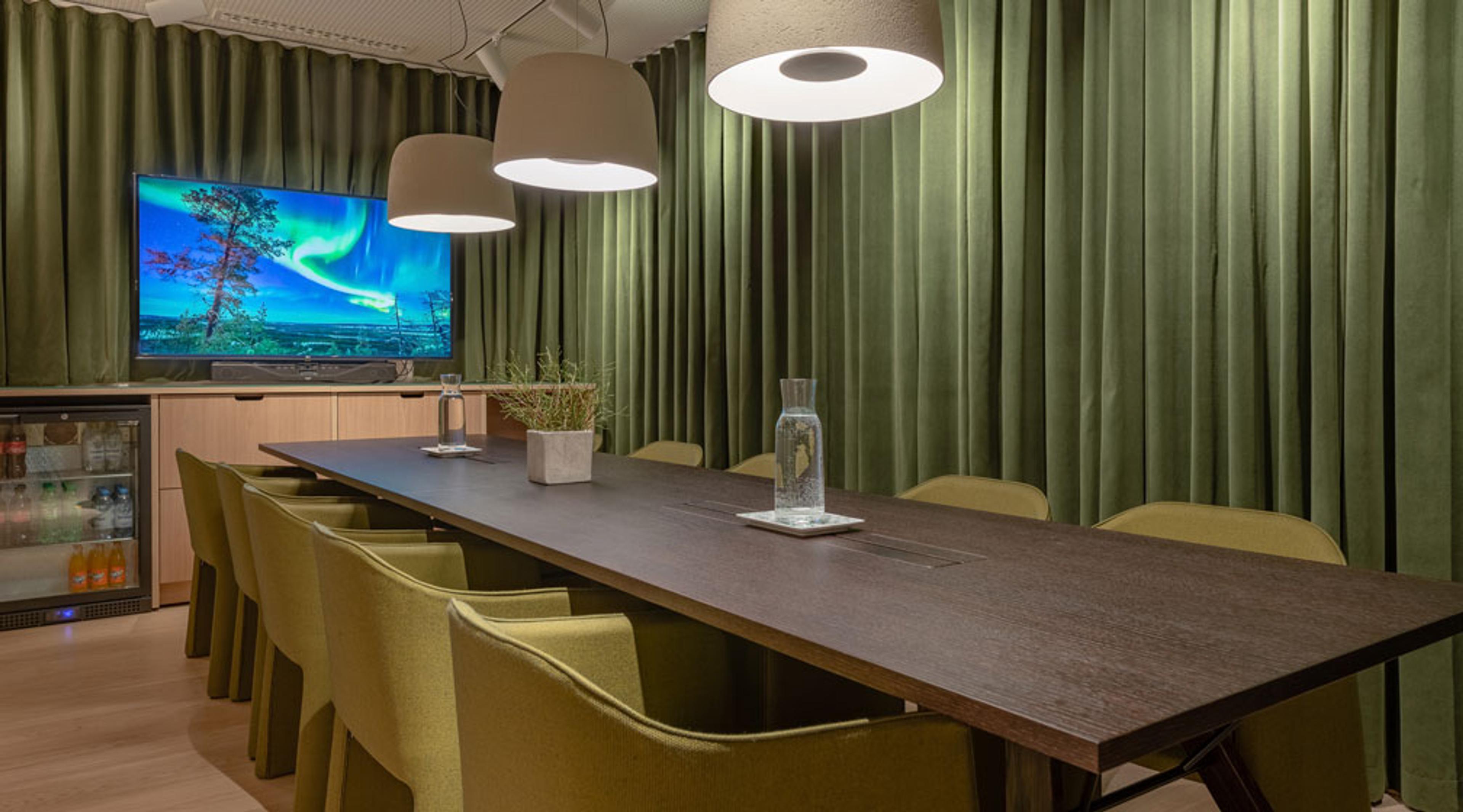 Moderni neuvottelutila vihreillä verhoilla, pitkä pöytä, tyylikkäät tuolit ja televisioruudulla revontulia. Vaaleat puulattiapinnat ja kattovalaisimet.
