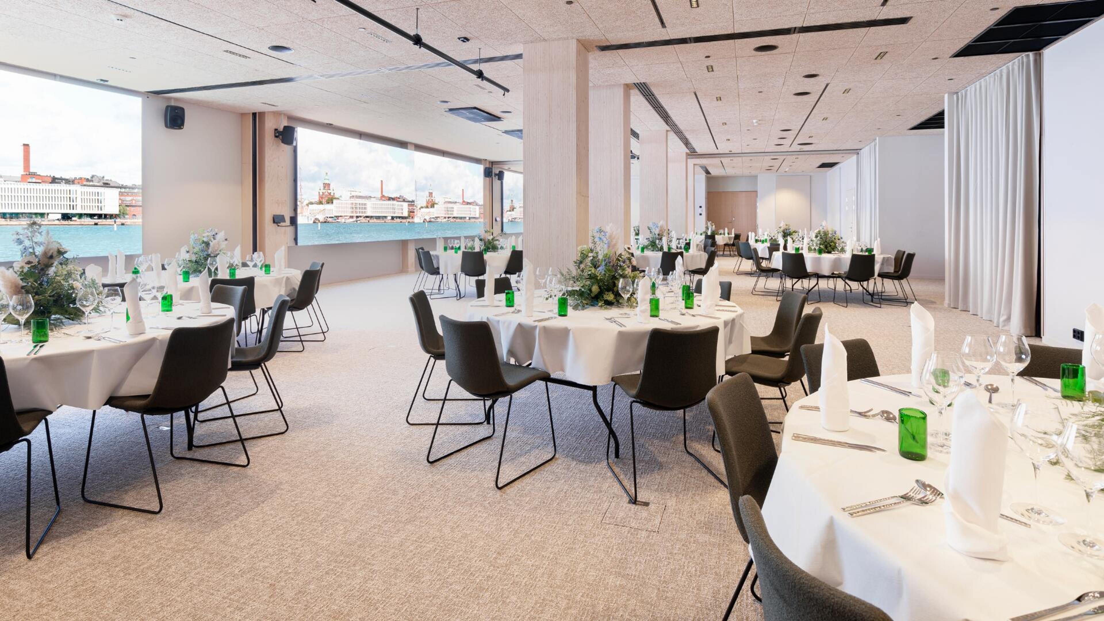 Moderni juhlatila pyöreiden pöytien kanssa. Suuresta ikkunasta avautuu näkymä kaupungin satamaan. Lattialla on matto ja tilassa on esteetön kulku pöytien välissä.