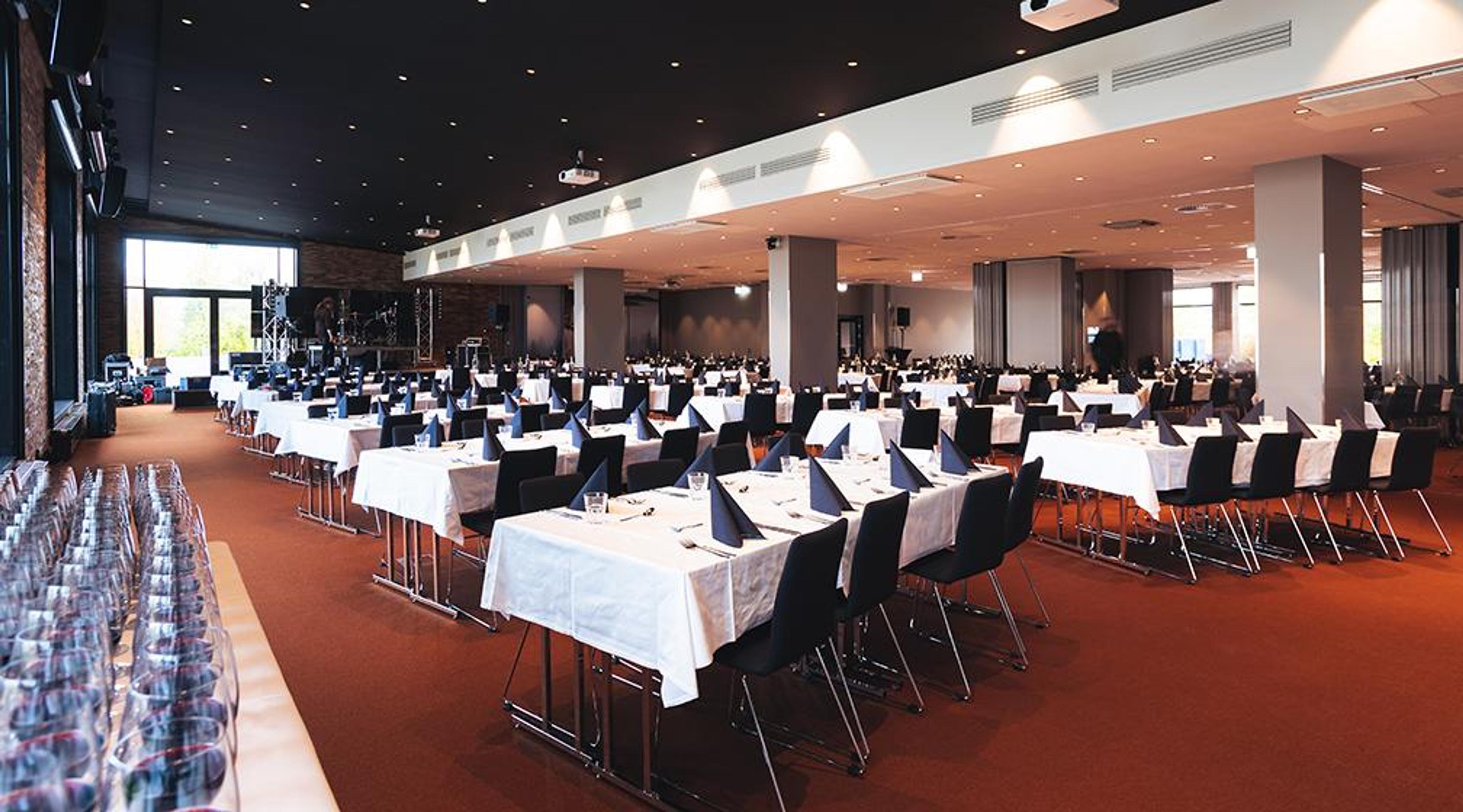 Spacious conference hall with white tablecloth-covered tables and black chairs. Stage area at the front with good overhead lighting. Red carpet flooring.