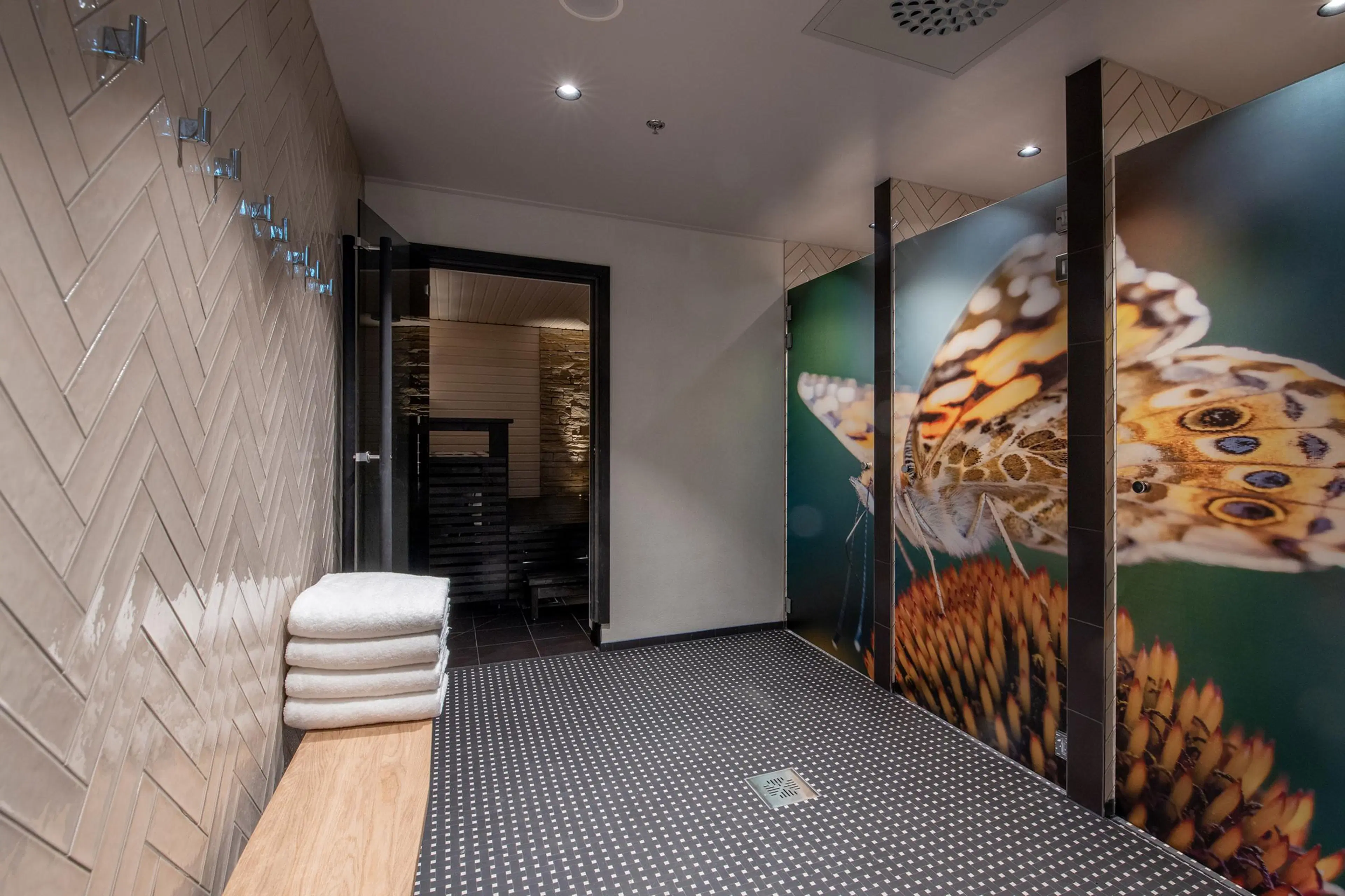 Saunan eteinen vaalein laatoitetuin seinin. Pinossa valkoisia pyyhkeitä puupenkillä, lattia tumma ja ovi saunaan auki. Oikealla perhoskuvioinen lasiseinä.