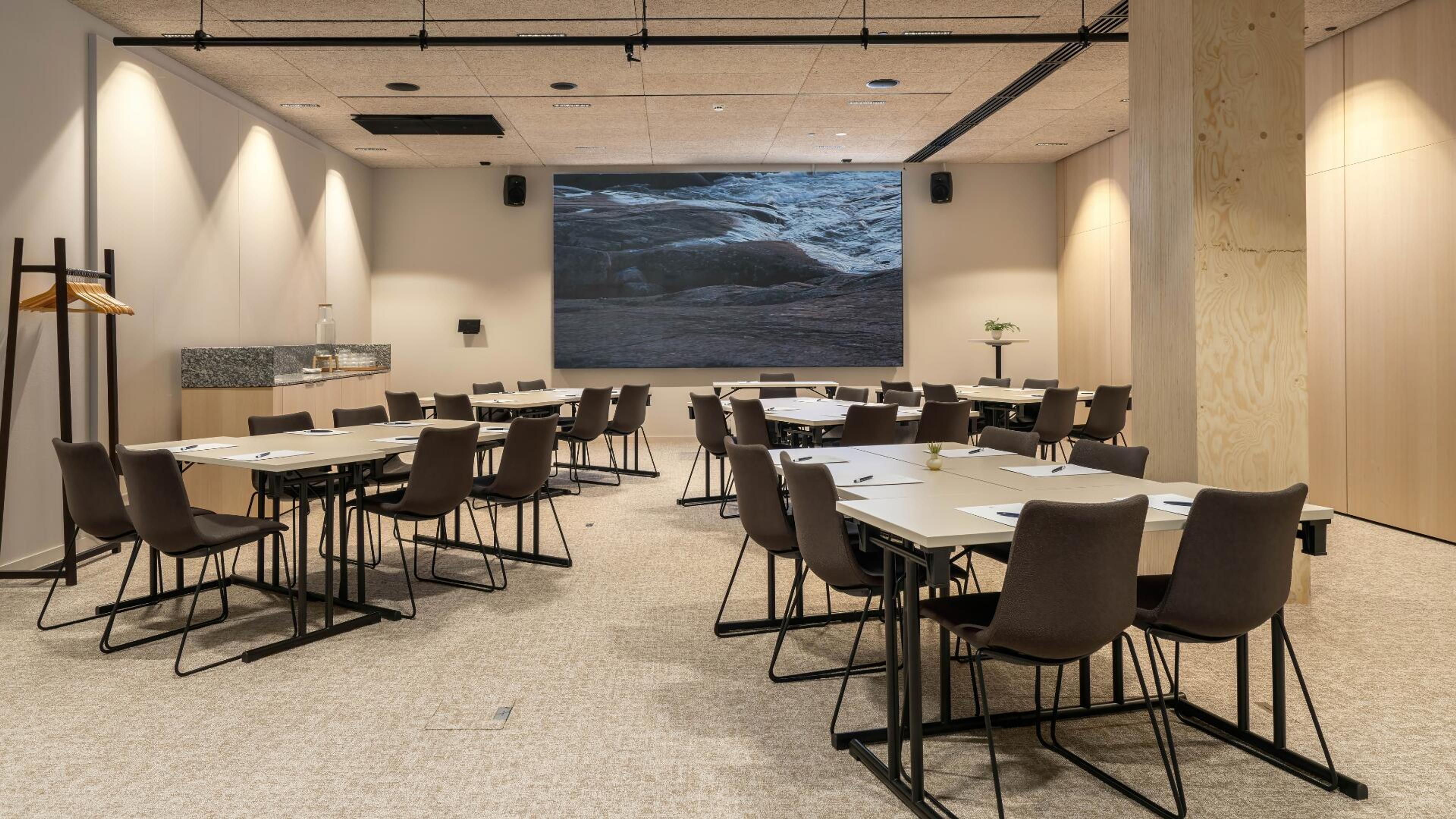 Moderni kokoustila, jossa pöytäryhmiä, ruskeat tuolit, suuri näyttö edessä ja tasainen kokolattiamatto. Seinillä vaaleat puupaneelit ja kohdevalaistus.