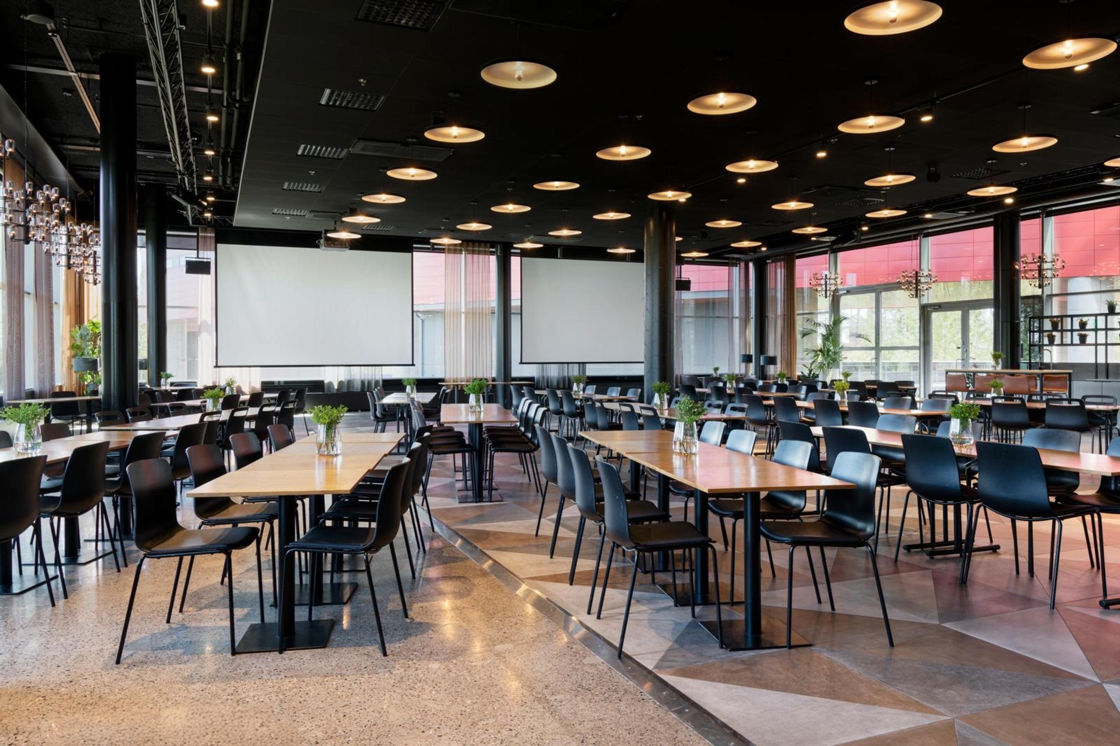 Modernt konferensrum med träbord och svarta stolar. Två projektionsdukar, plant stengolv och stora fönster. Taklampor skapar en varm atmosfär.