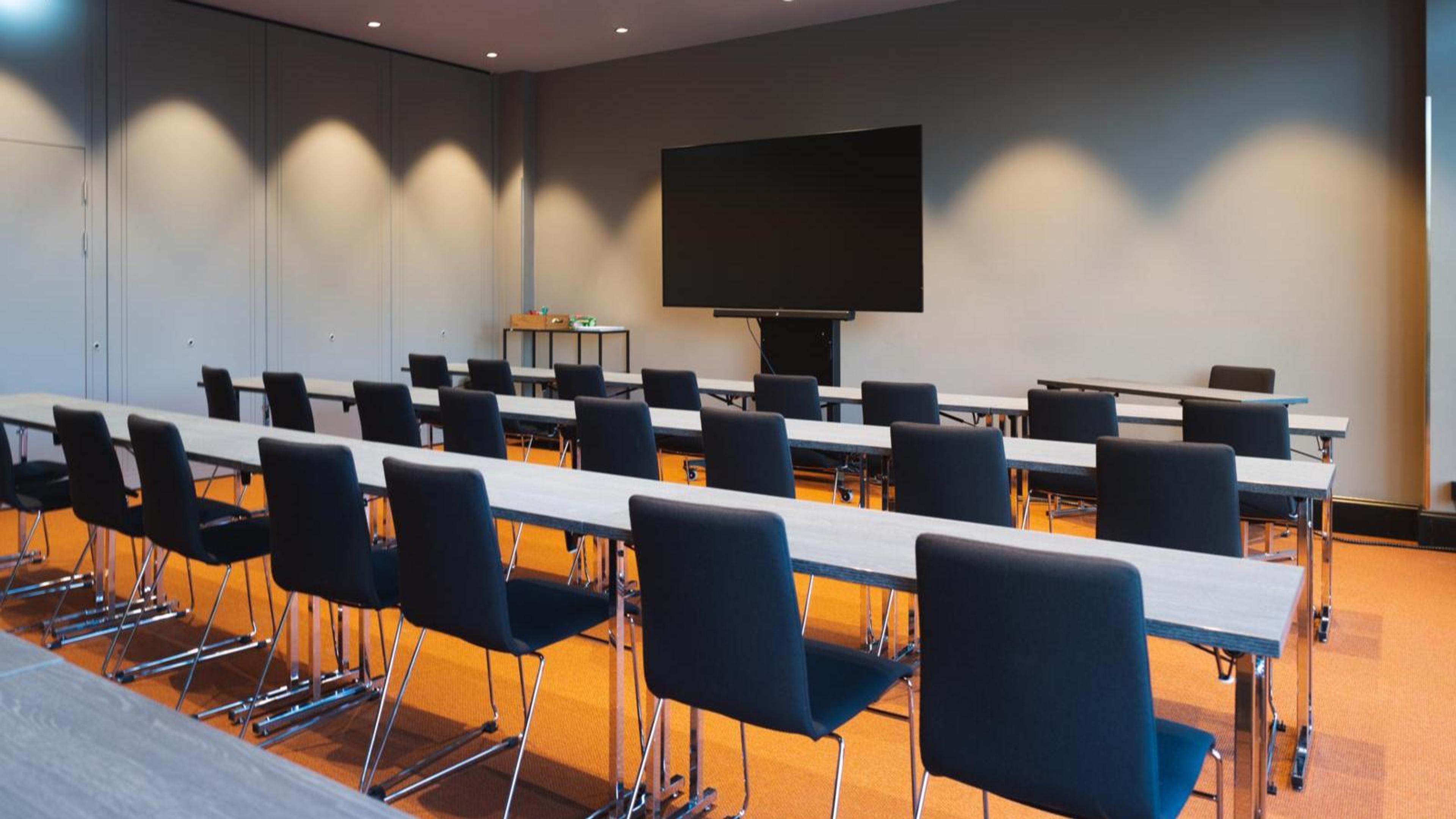 Conference room with long tables and black chairs arranged in rows, large screen at the front. Room has level flooring and good lighting.