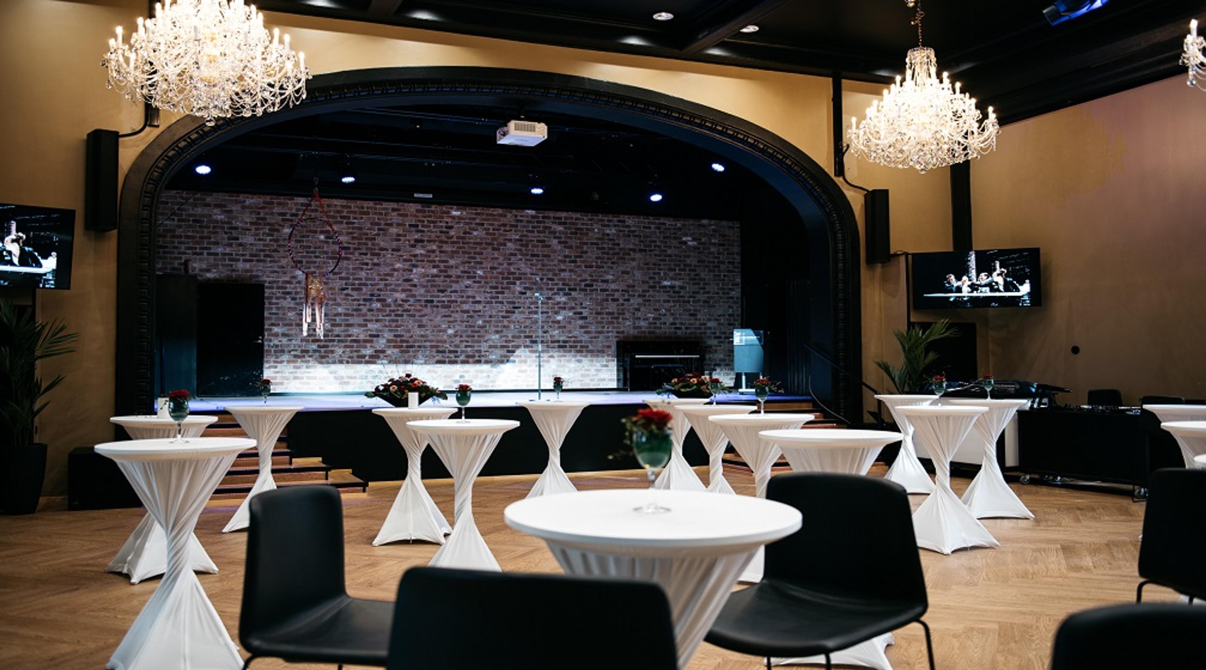 Juhlasali, jossa on valkoisia cocktail-pöytiä, elegantteja kristallikruunuja, puulattia ja tiiliseinäinen esiintymislava.
