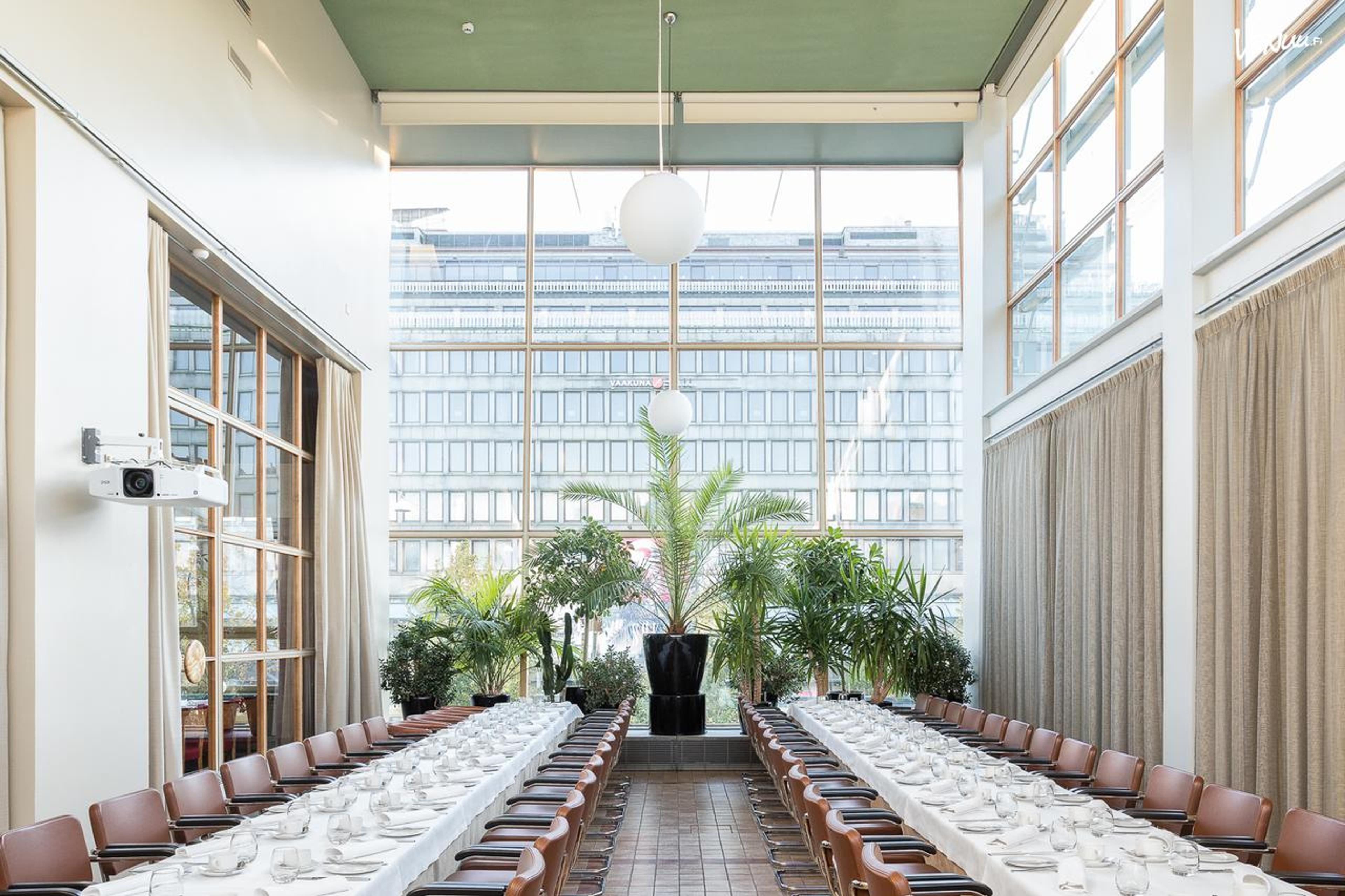 Valoisa tilava ravintolasali kahdella pitkällä pöydällä. Taustalla isot ikkunat kaupunkimaisemaan ja keskellä tilaa viherkasveja.