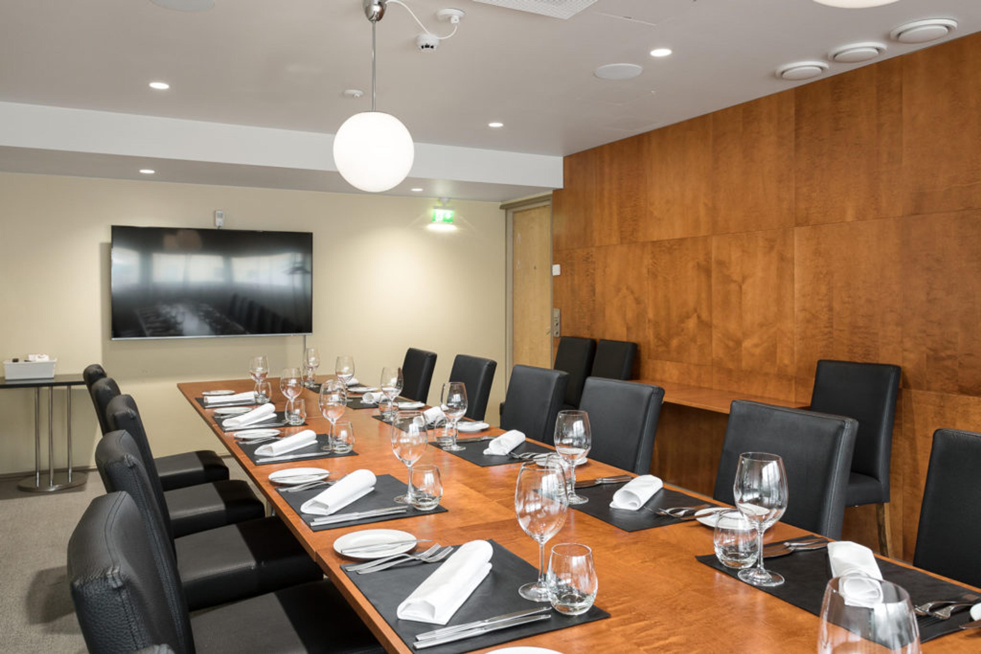 Kokoustila, jossa pitkä puinen pöytä, mustia tuoleja, kattava kattaus laseineen ja lautasliinoineen, seinällä iso näyttö ja puinen seinäpaneeli.