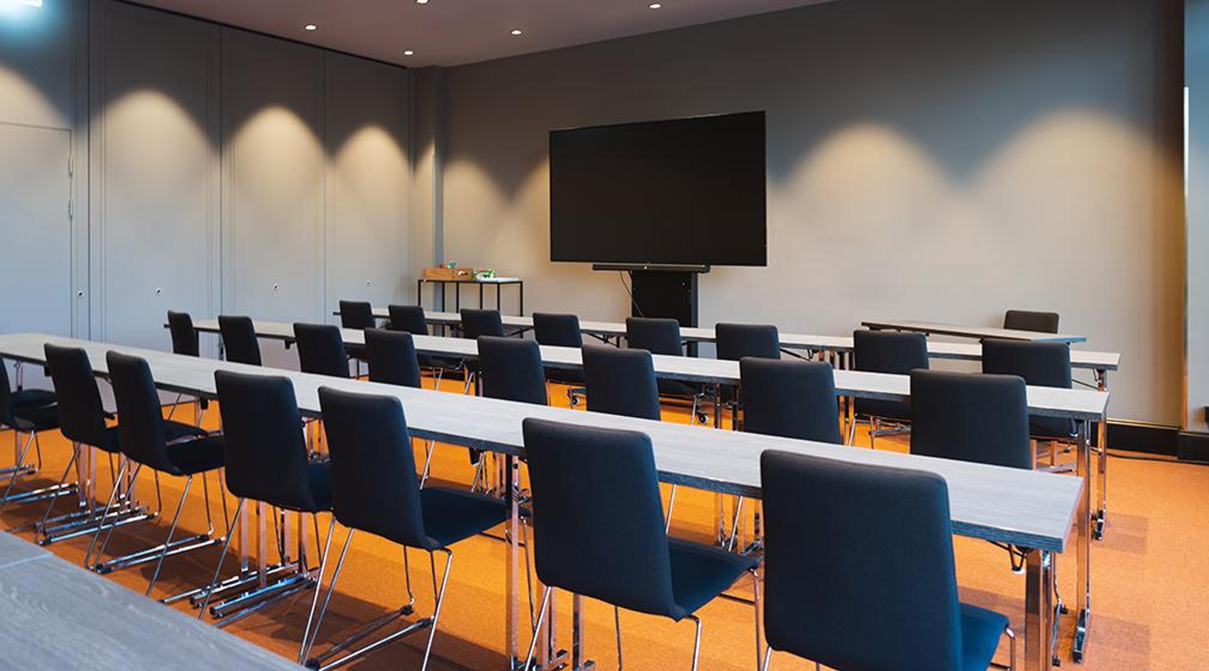 Modern conference room with rows of tables and blue chairs. Large display screen at front. Room has spotlights and orange carpeting.