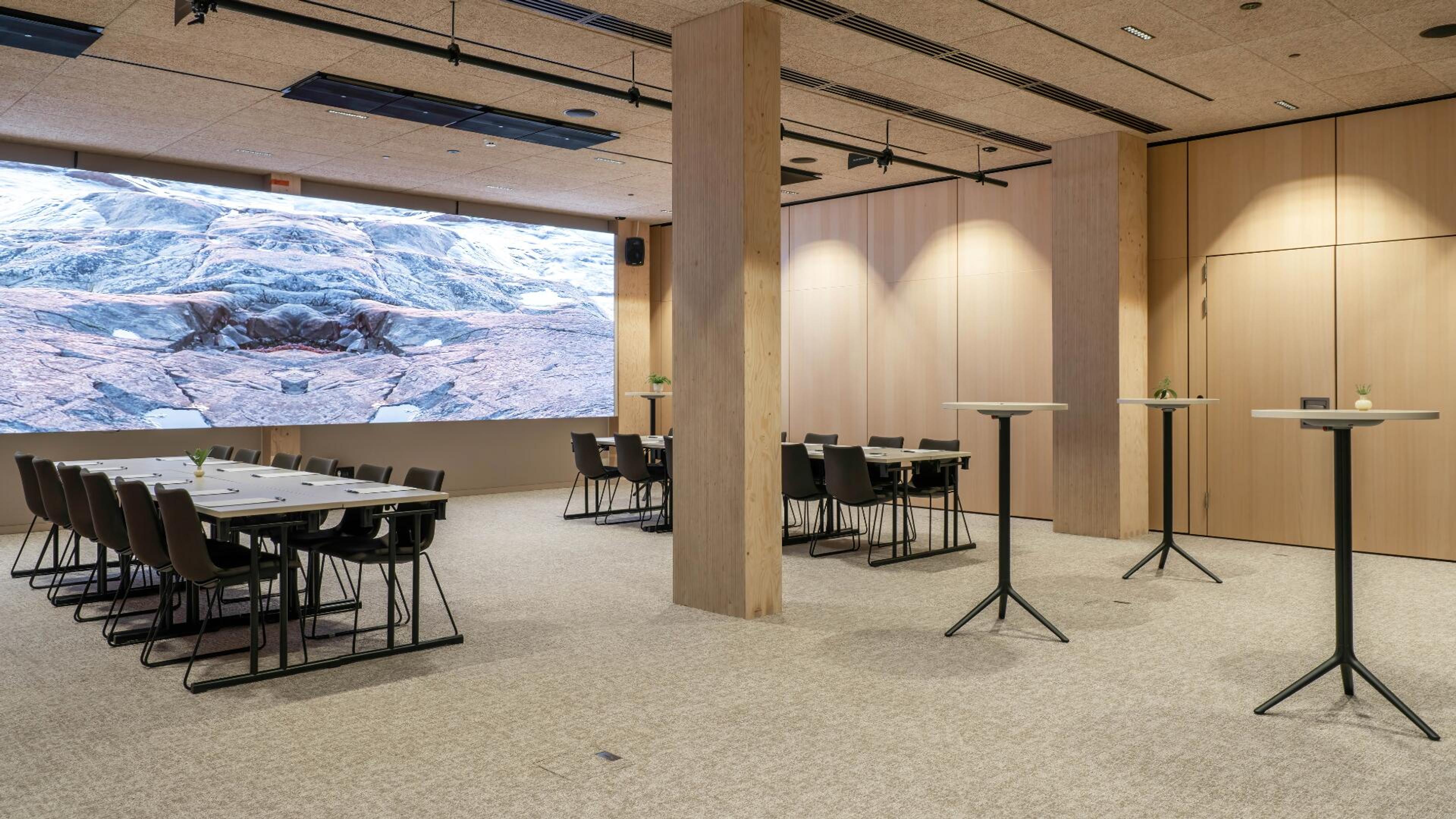 Moderni kokoustila puisilla yksityiskohdilla, suurella näytöllä, neuvottelupöydillä ja korkeilla cocktailpöydillä. Tasainen lattia ja avara tila.