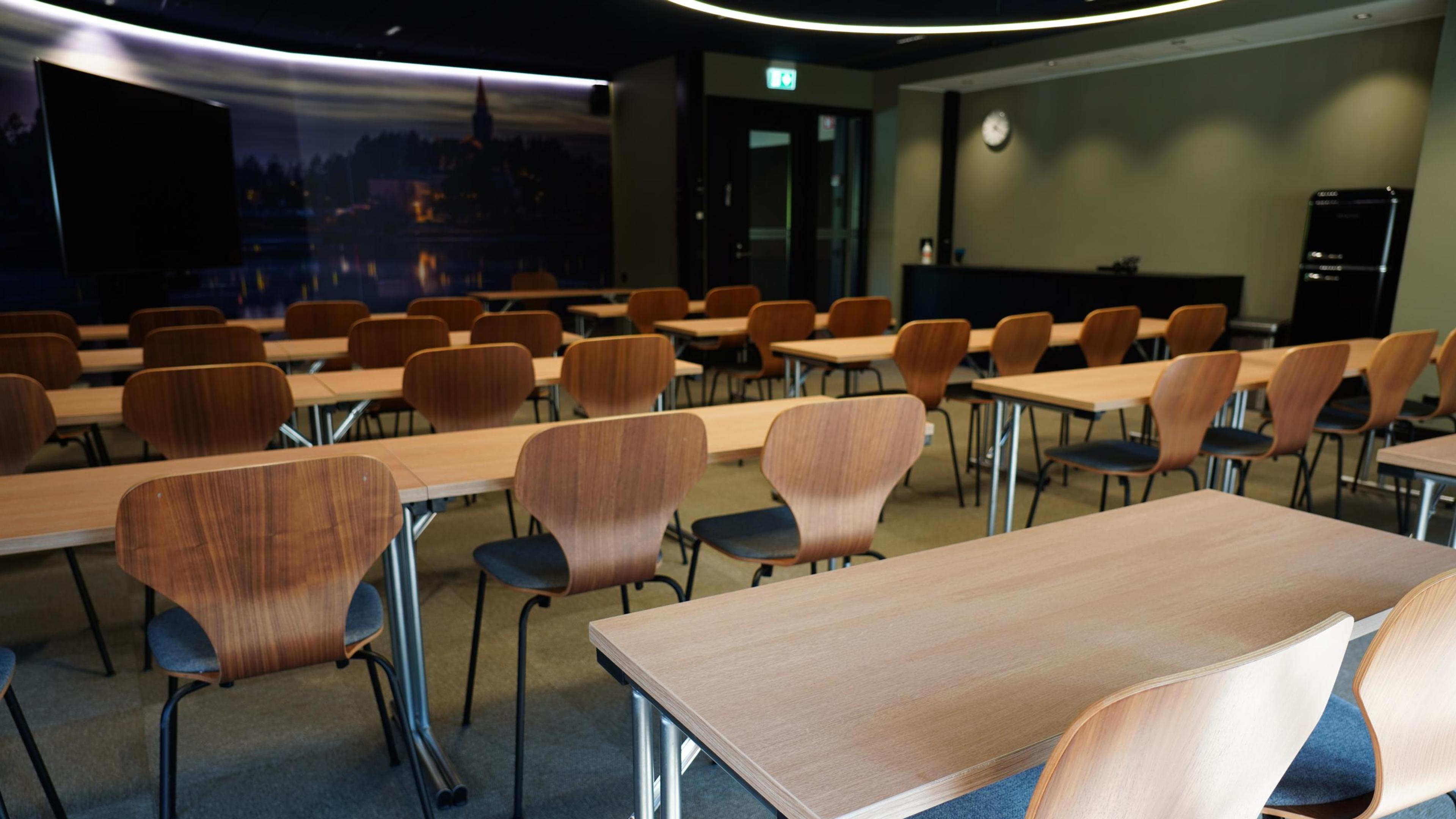 Ett tomt mötesrum med bord i rader och trästolar. En stadsbild på väggen och en bred dörr i bakre delen.