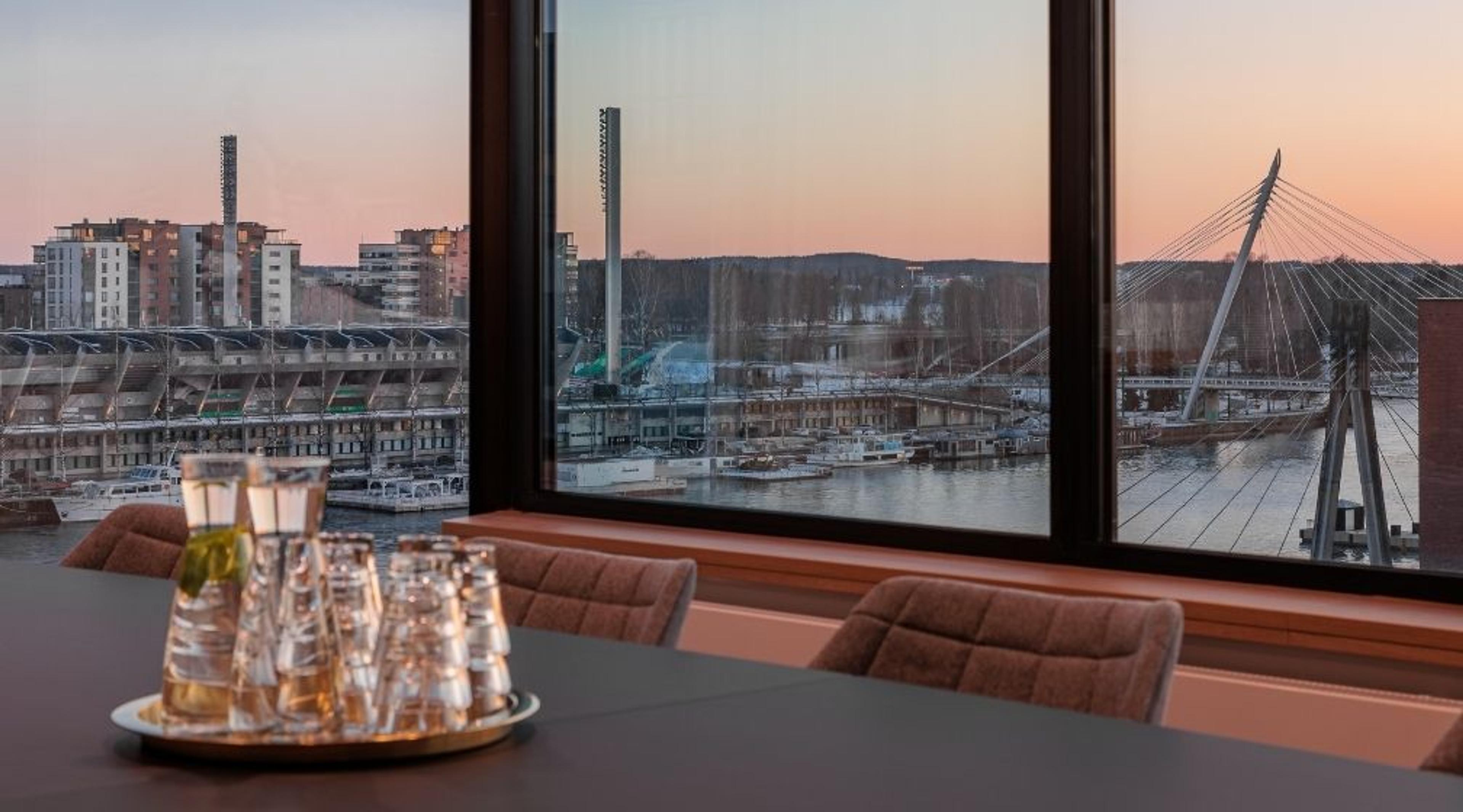 Kokoushuone, jossa on iso ikkuna. Lasit tarjottimella pöydällä. Näkymä joelle, stadionille ja siltaan auringonlaskun aikaan.