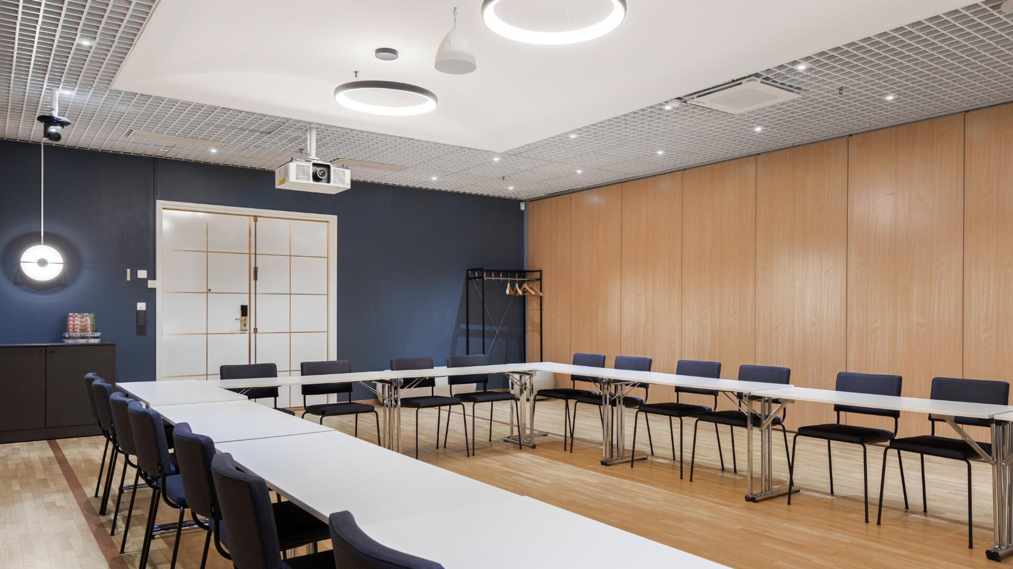 Valoisa kokoustila, jossa pöydät on järjestetty U-muotoon, sinisiä tuoleja, puinen lattia, puupaneeliseinä ja sininen seinä, kattovalaisimet ja projektori.