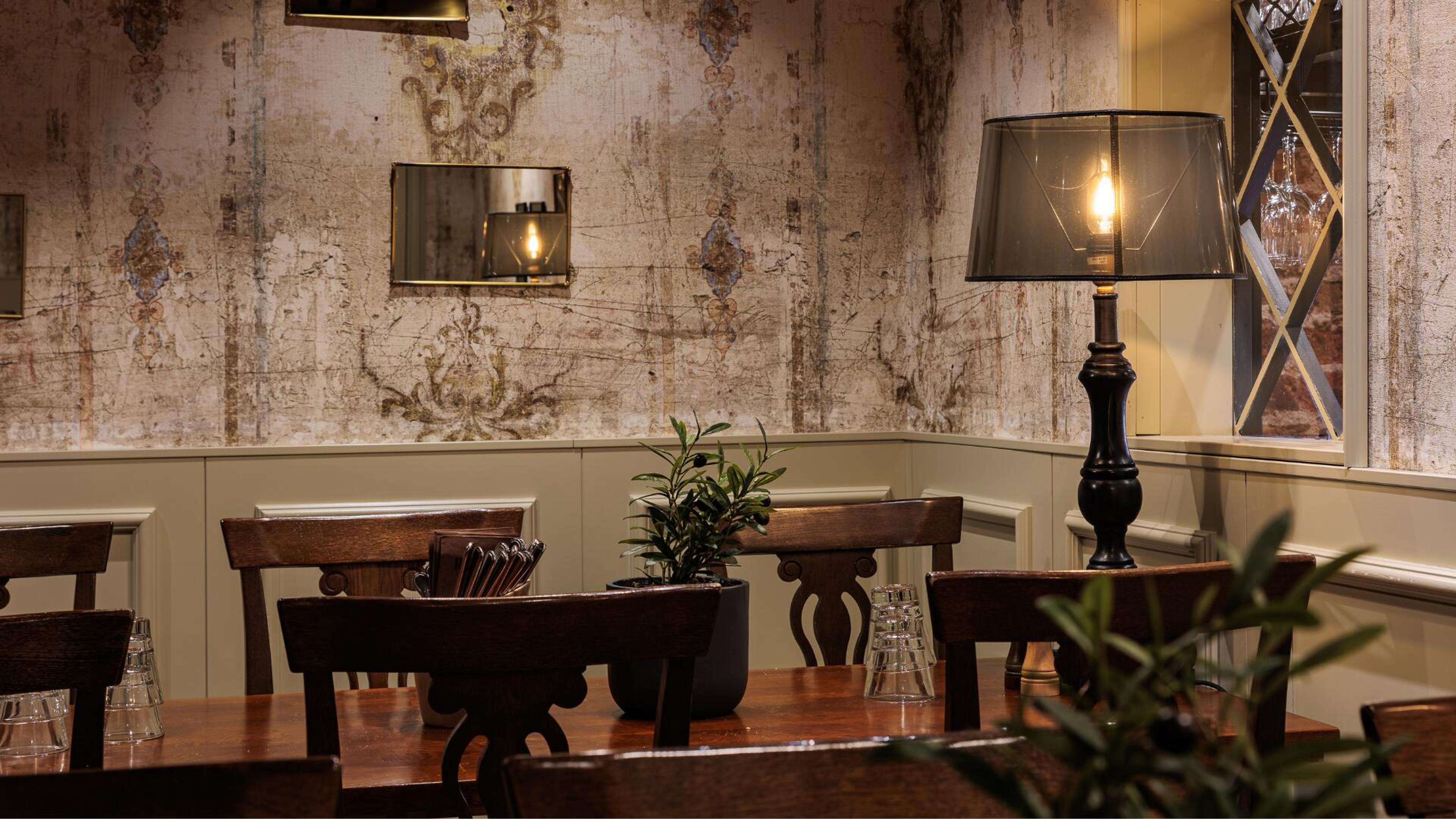 Ravintolan kabinetti, jossa tunnelmallinen sisustus, jossa puisia pöytiä ja tuoleja, ornamenttikuvioitu tapettiseinä ja musta pöytävalaisin.