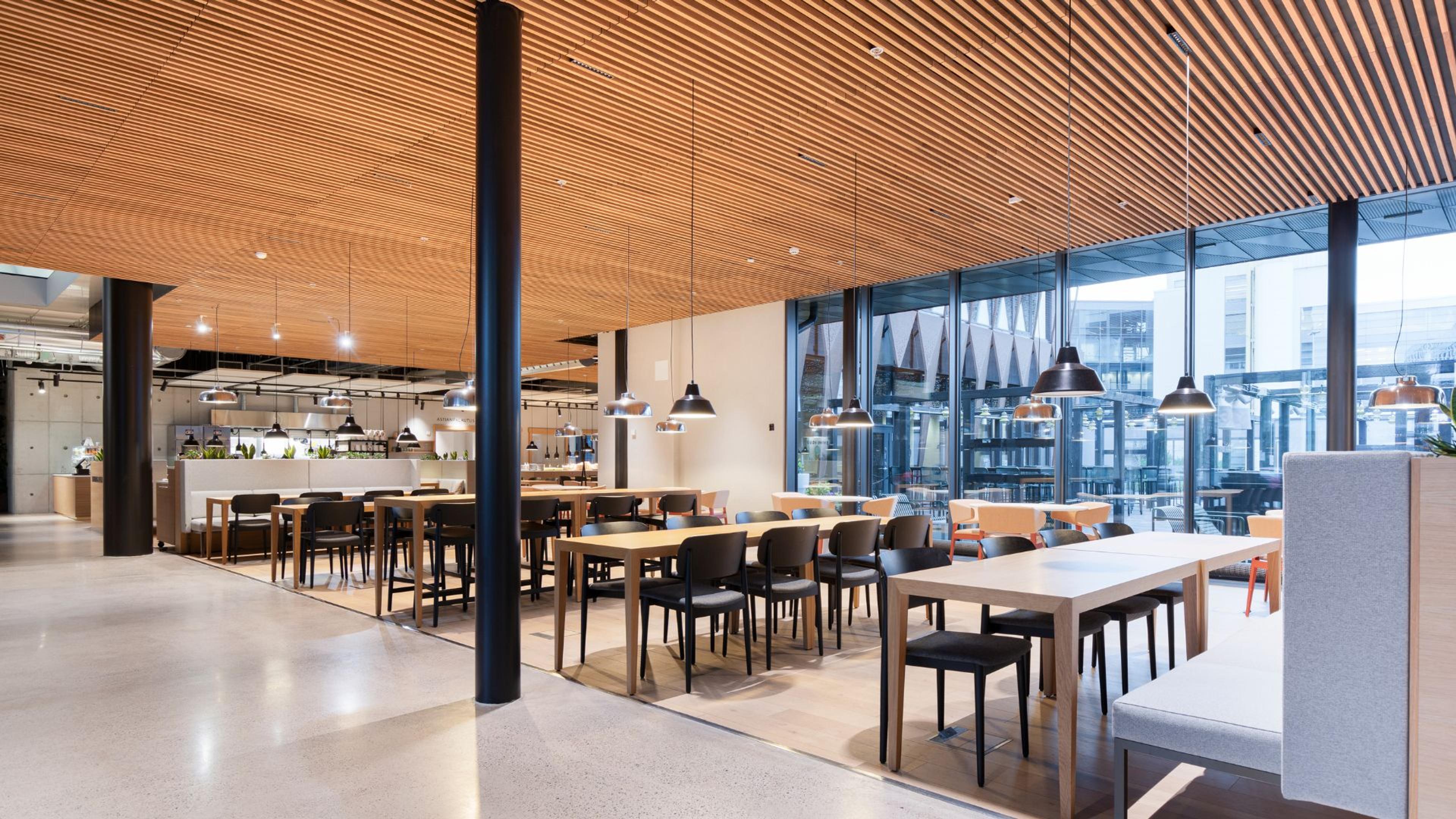 Moderni ravintola puisella kattopaneloinnilla, avoimella pohjalla ja isoilla ikkunoilla. Tasainen lattia ja leveät kulkuväylät pöytien välissä.