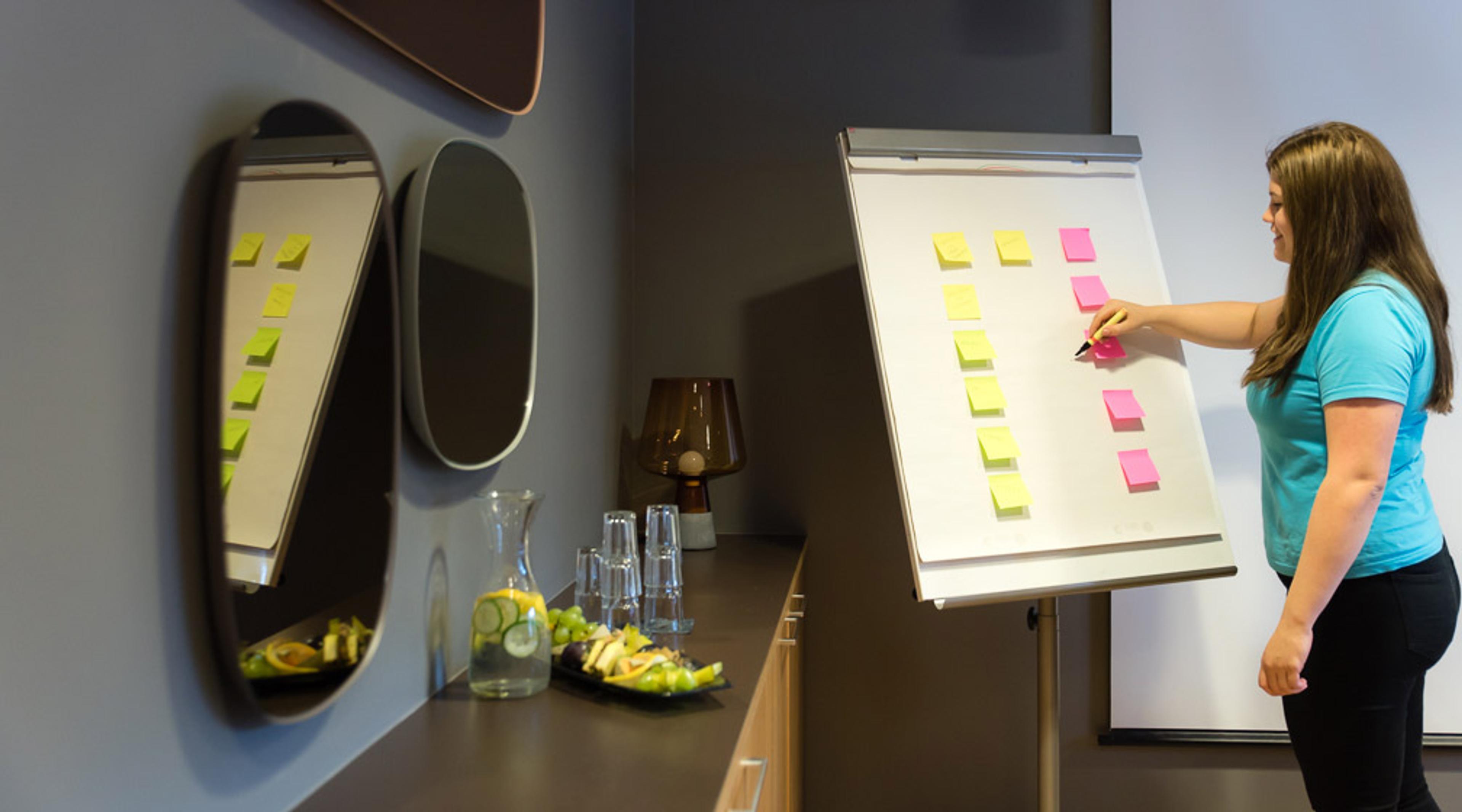 Person in blue shirt writing on sticky notes on a flipchart. A carafe and water glasses sit on the counter in the background.