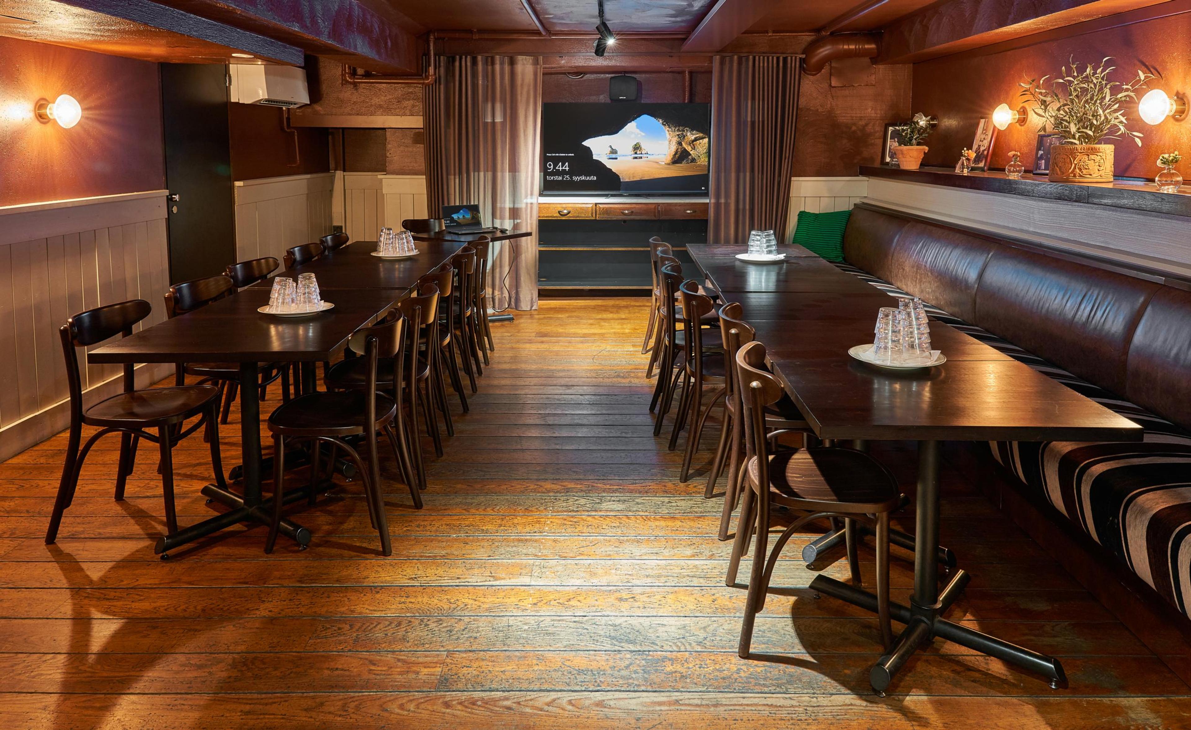 Kokoustila puisella lattialla, jossa kaksi pitkää pöytää, tuolit ja sohva. Taustalla näyttö, puiset seinäpaneelit ja lämpimät valot.