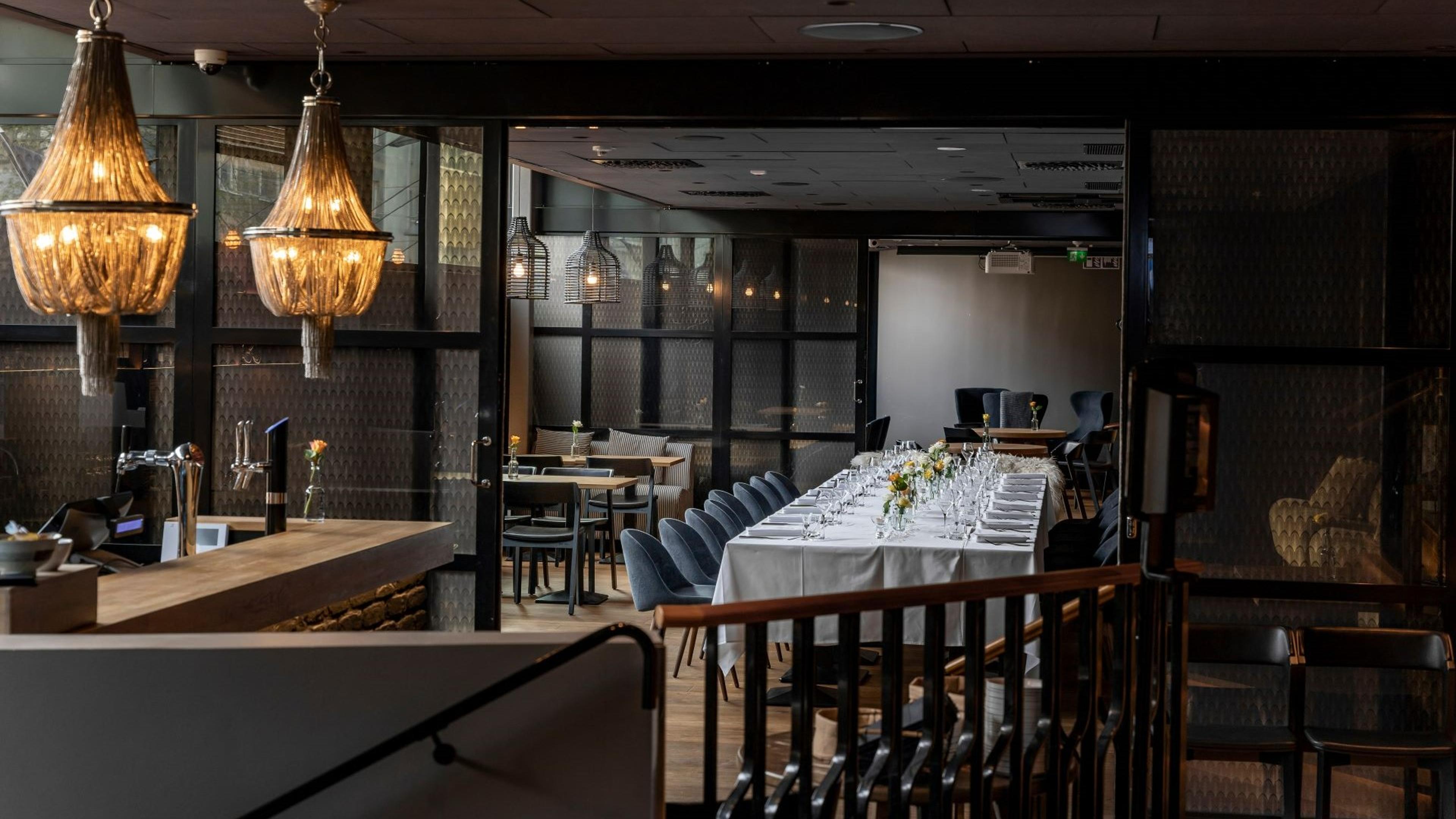 Ravintolasali, jossa elegantti kattokruunu, baaritiski vasemmalla ja pitkä juhlapöytä valkoisella liinalla ja sinisillä tuoleilla. Puulattia ja lasiseinät.