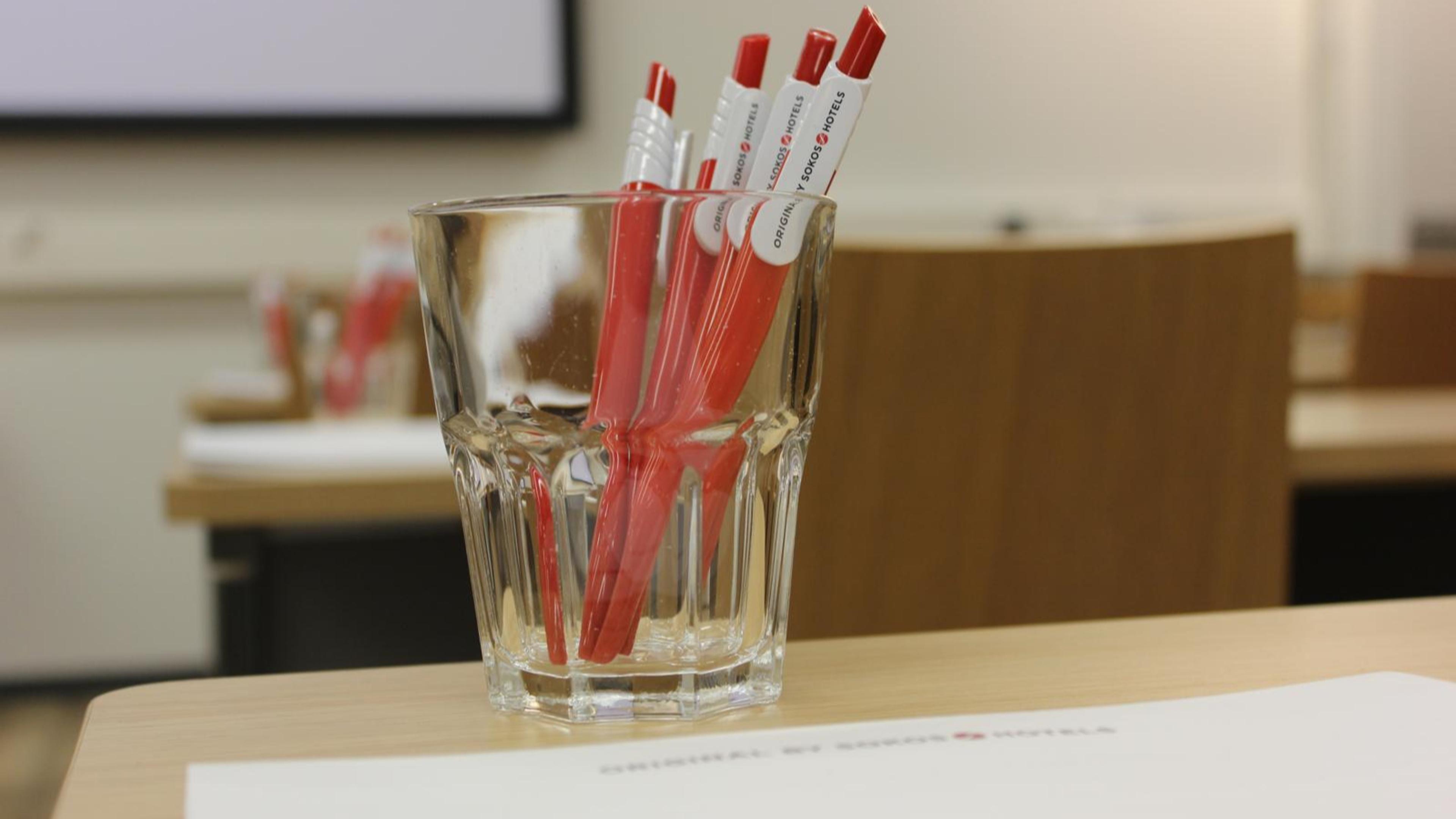 Röda pennor i ett glasigt kärl på ett träbord, med konferensrumsstolar och en skärm i bakgrunden.