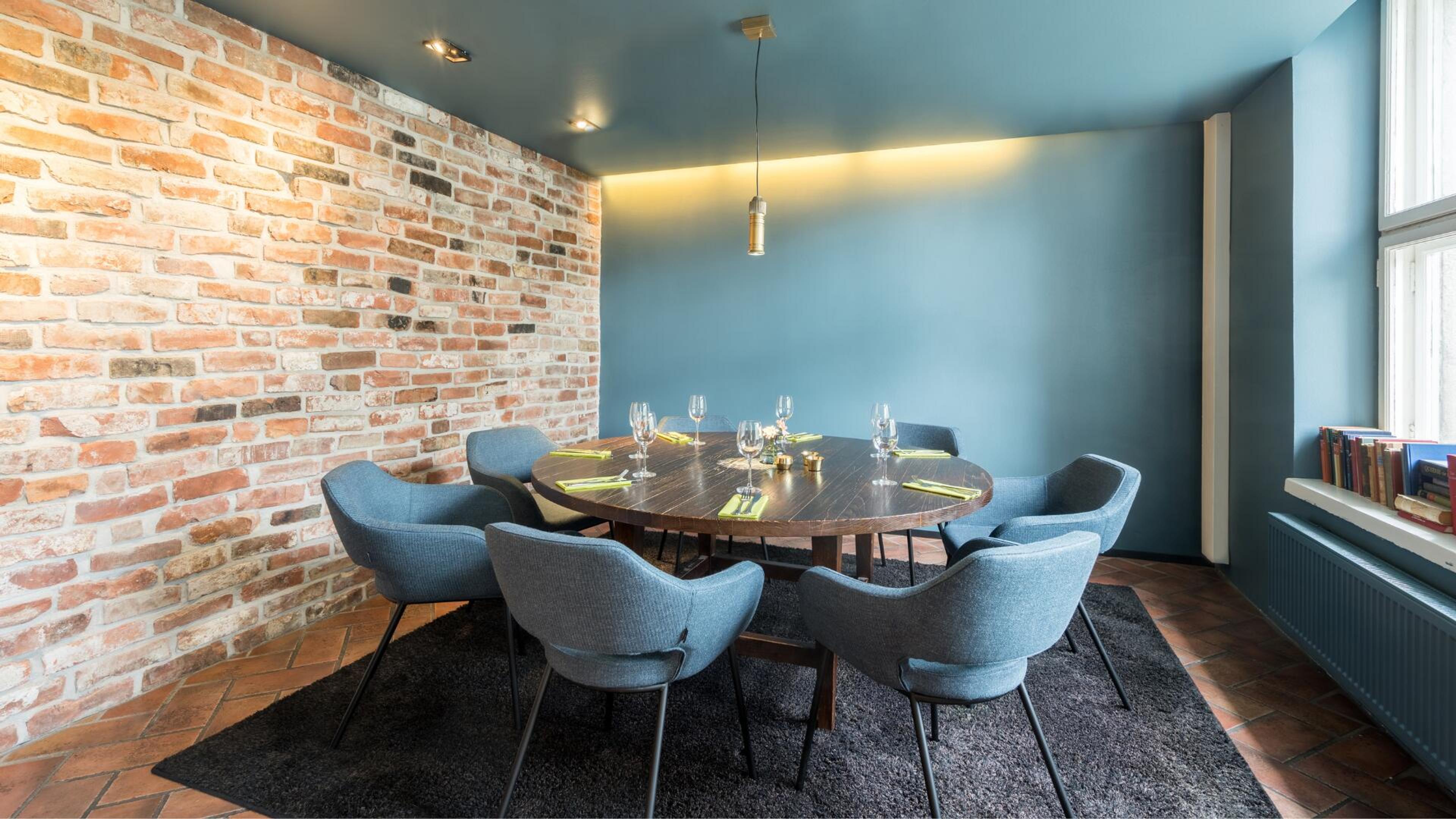 Yksityinen ruokailutila, jossa pyöreä puupöytä, sinisiä nojatuoleja, tiiliseinä ja sininen seinä. Tasainen laattalattia tummalla matolla.