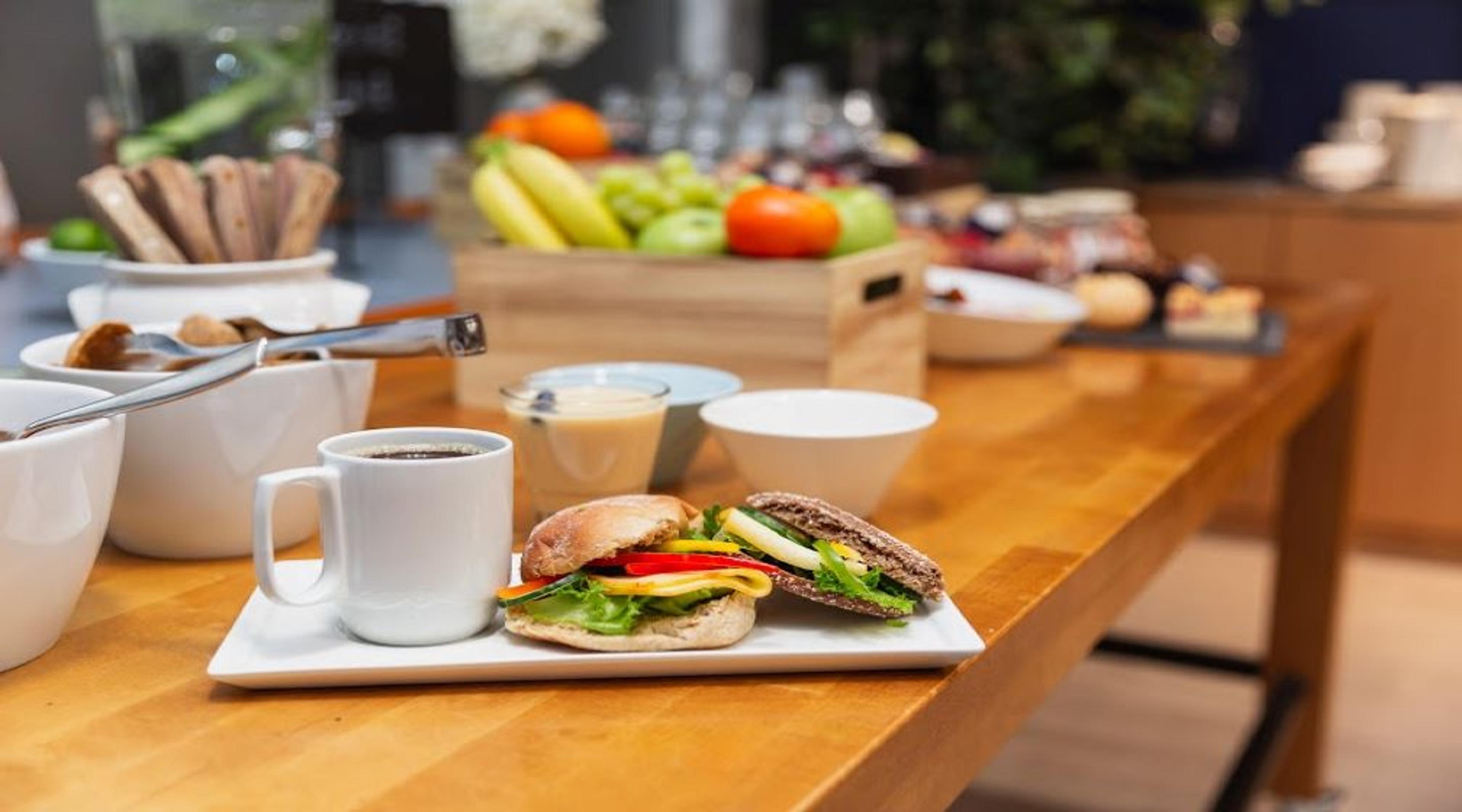Café breakfast on wooden table: filled bagel sandwich, coffee cup, and fruit basket with other delicacies in the background.