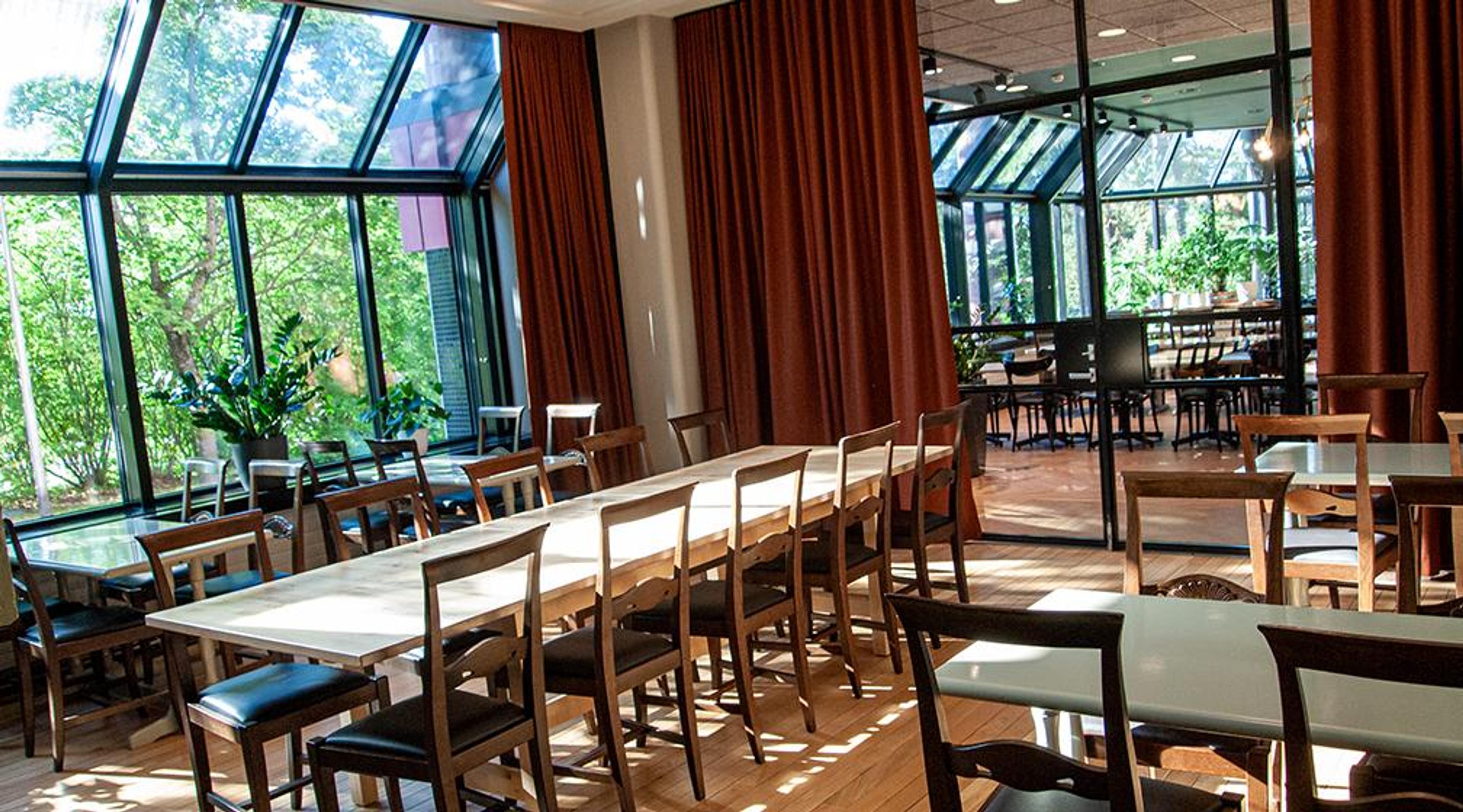 Ljus restaurang med långt träbord och stolar, glasväggar med trädgårdsutsikt och rödbruna gardiner. Trägolv och ytterligare utrymme syns bakom glasdörrar.