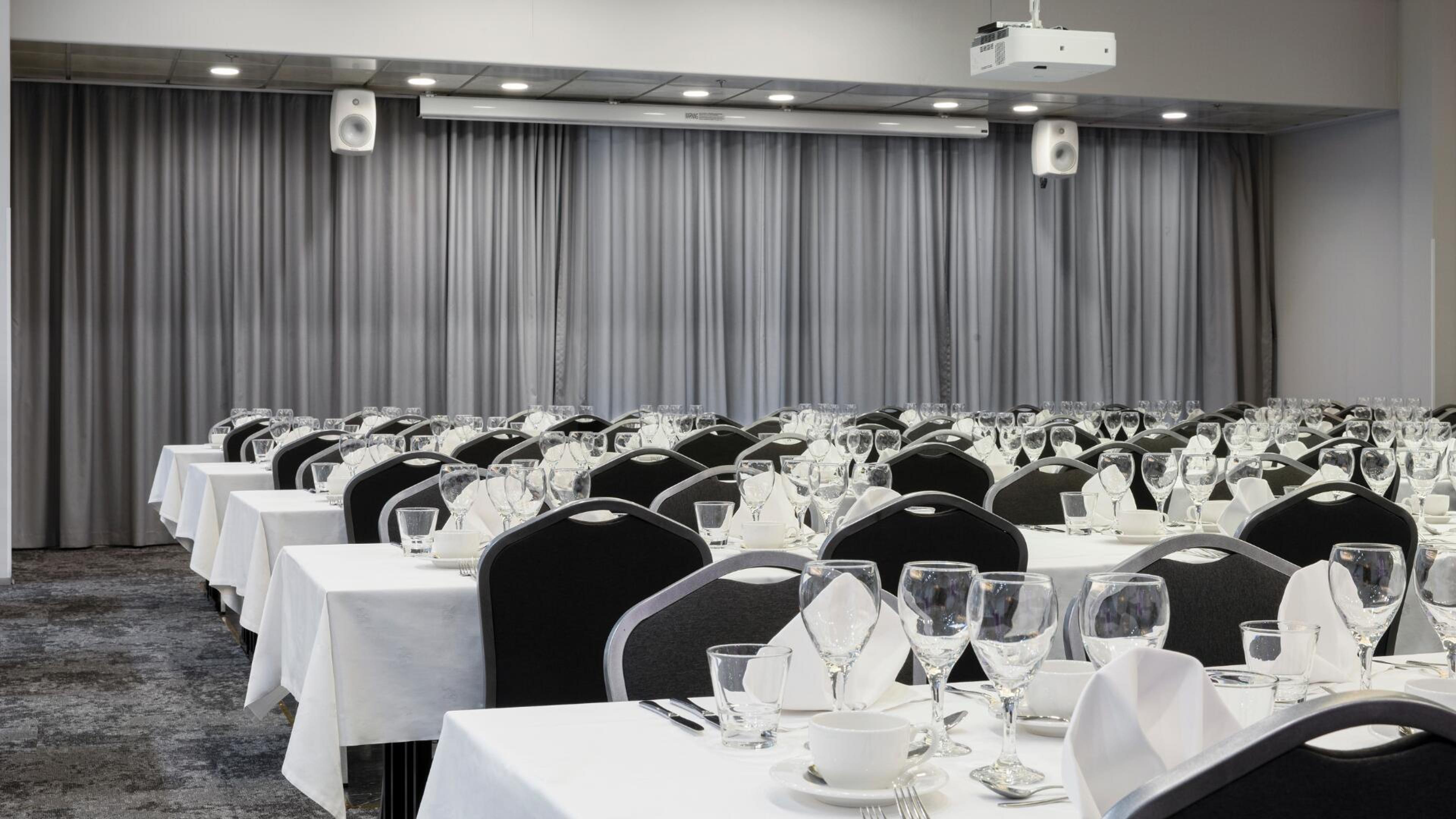 Kokoustila, jossa pöydät valmiina illallista varten. Valkoiset pöytäliinat, viinilasit ja astiasto. Mustia tuoleja ja harmaat verhot takaseinällä.