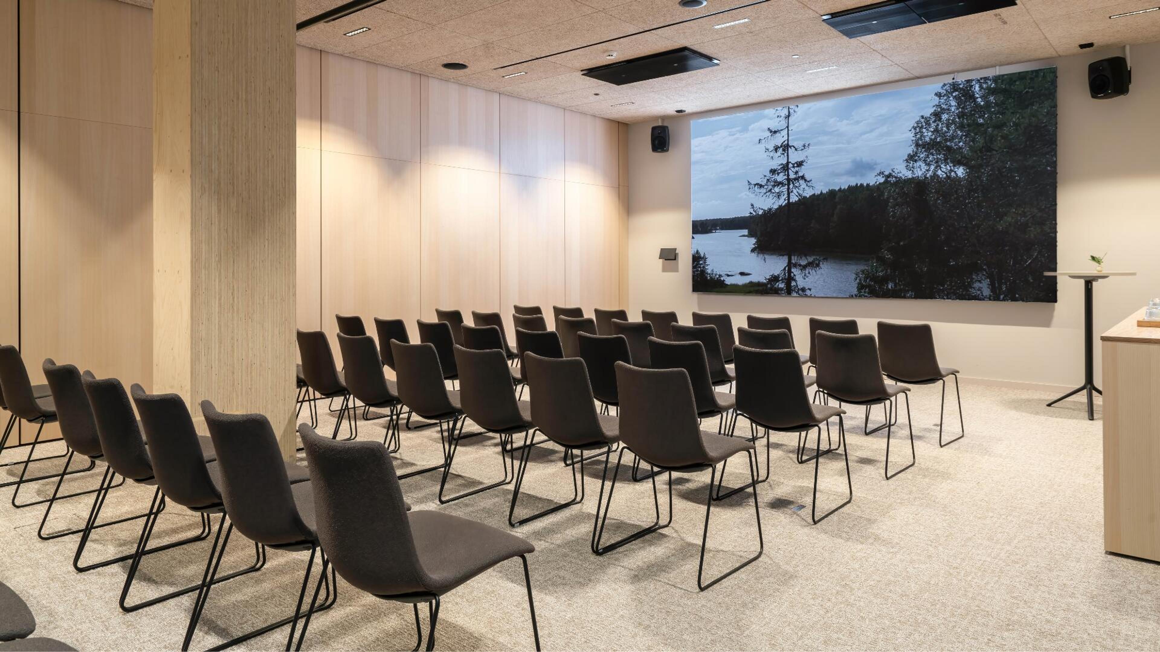 Moderni auditoriotila järjestetty tuoleilla riveihin. Seinällä suuri screeni, jossa on luontokuva järvimaisemasta. Vaalea lattia ja puupaneloidut seinät.