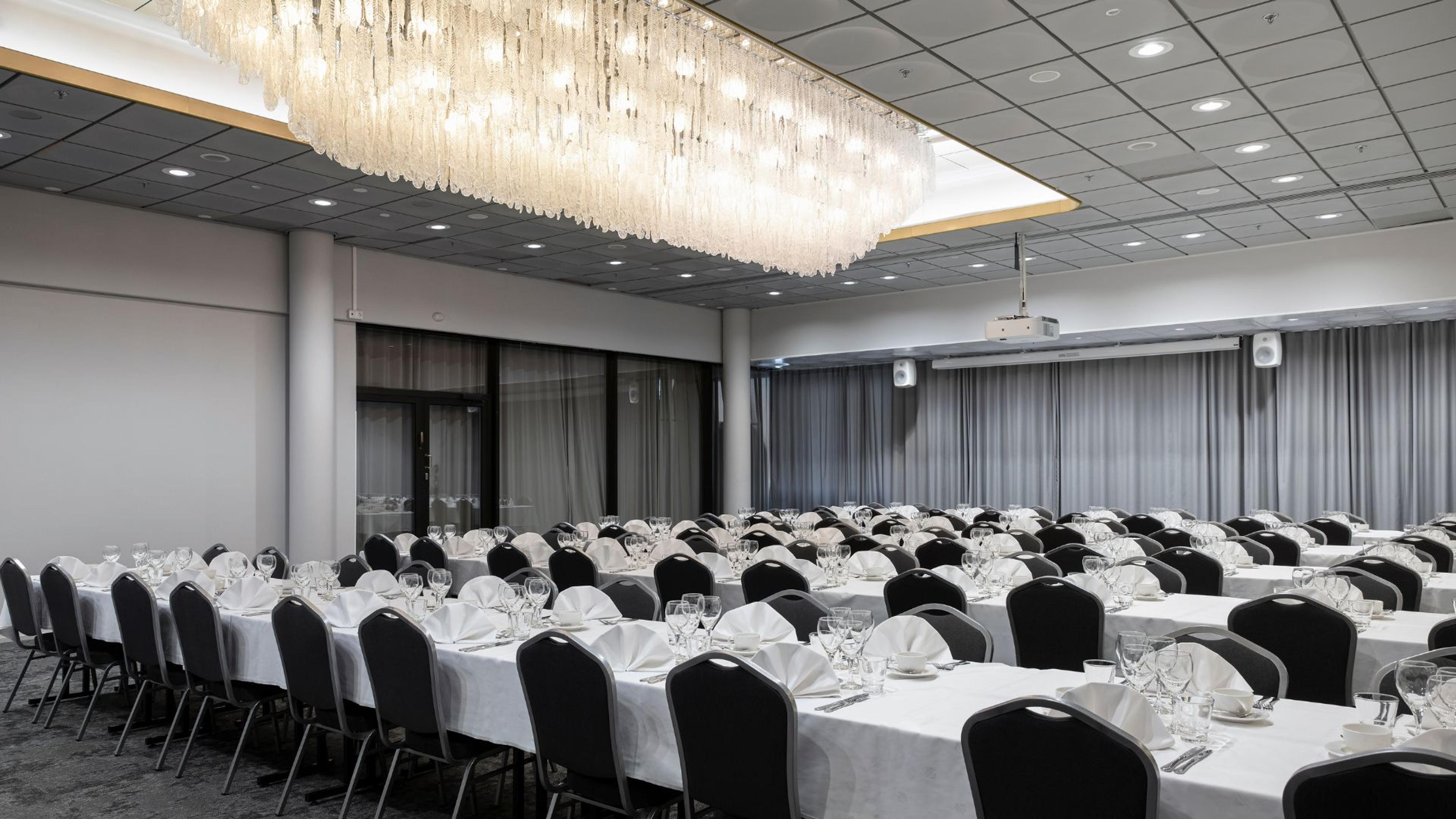 Elegantti juhlasali, jossa pitkät valkoisilla liinoilla katetut pöydät. Katossa näyttävä kristallikruunu, takaseinällä harmaat verhot ja projektori.