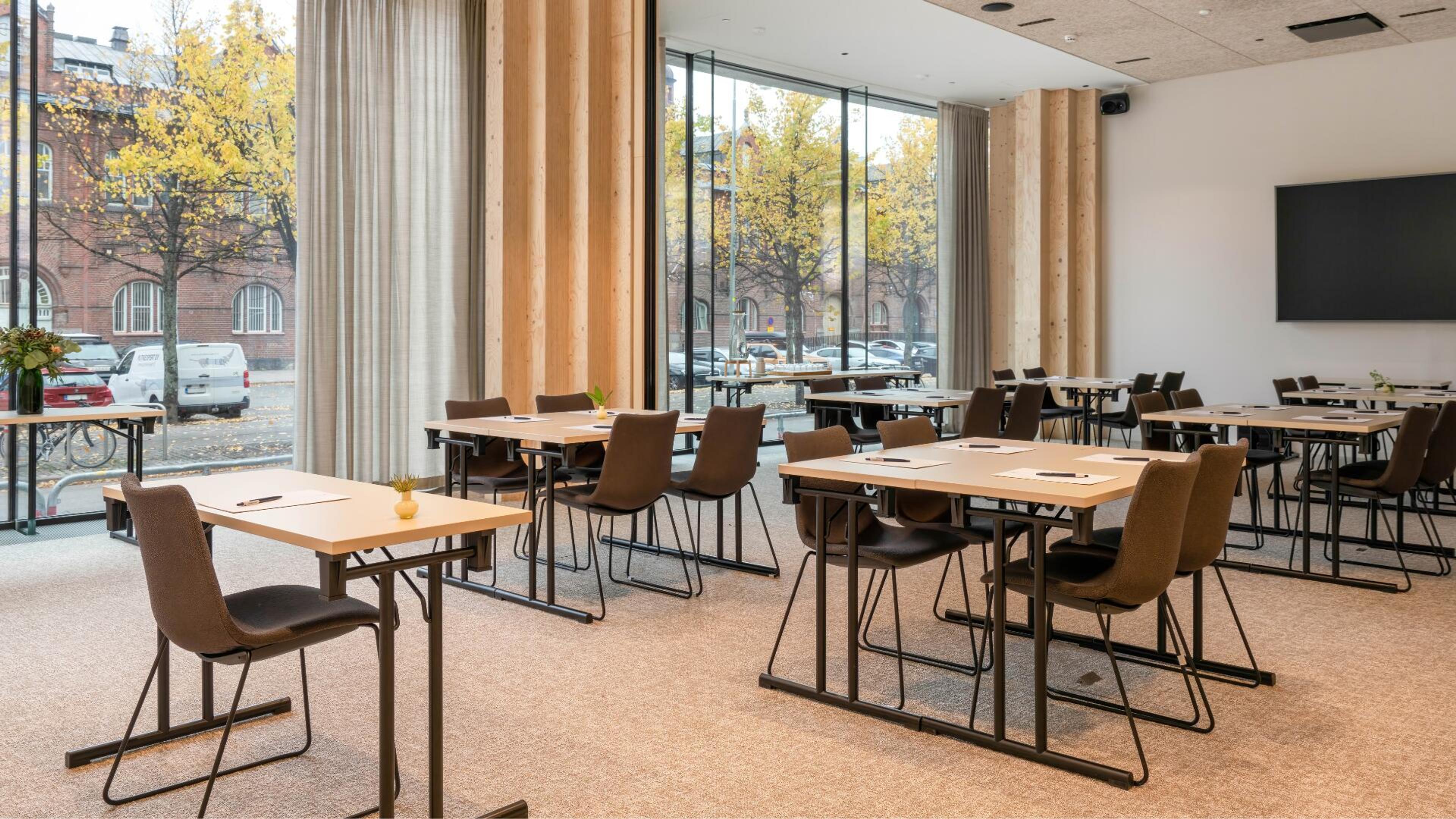 Moderni kokoustila puisilla pöydillä ja ruskeilla tuoleilla. Isot ikkunat tuovat luonnonvaloa ja näkymän syksyisille puille. Seinällä näyttö, lattia tasainen.