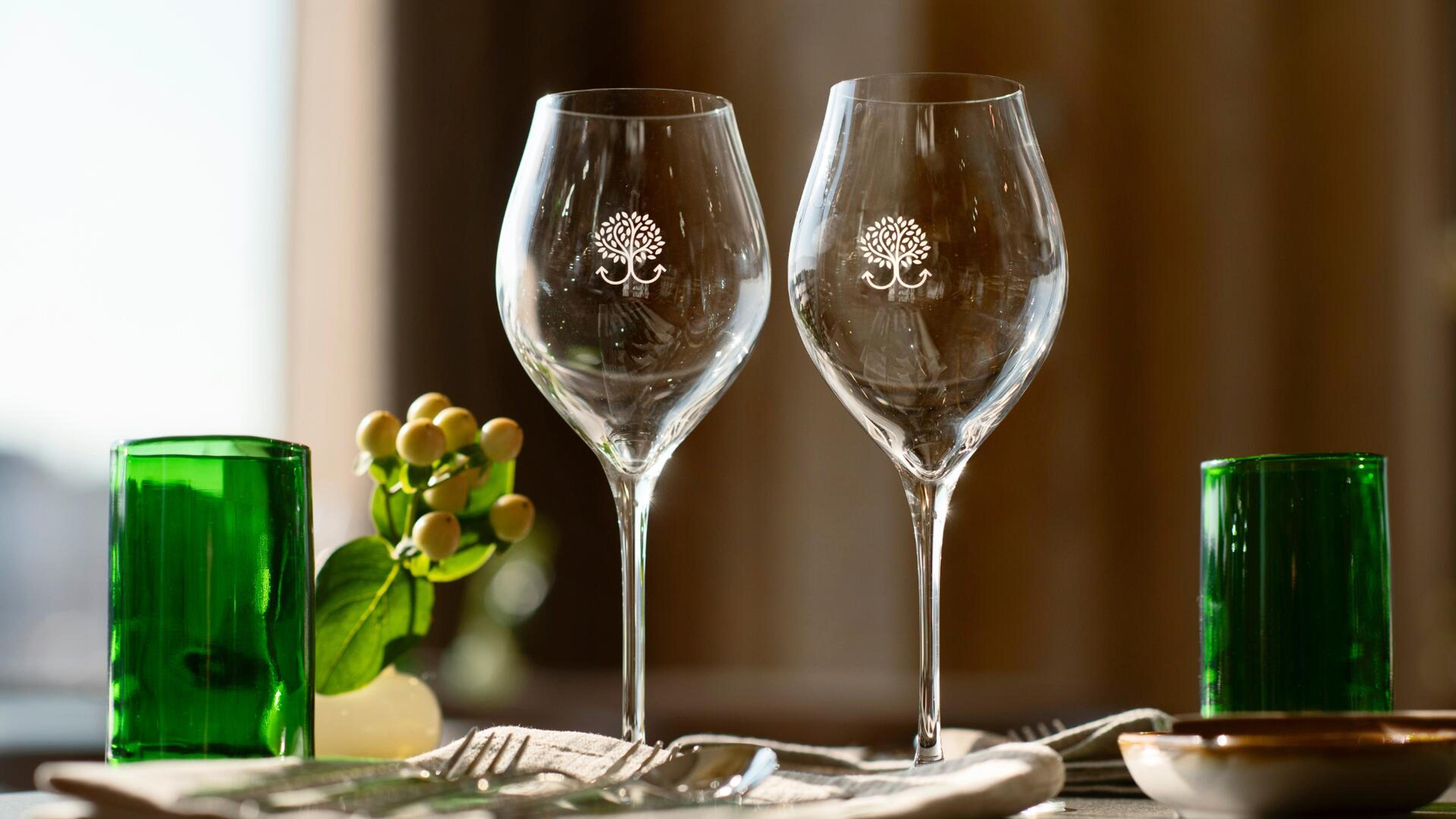 Kaksi tyhjää viinilasia, joissa on puu-logo, vihreitä juomalaseja ja pieni kasvi pöydällä aterimien kanssa.
