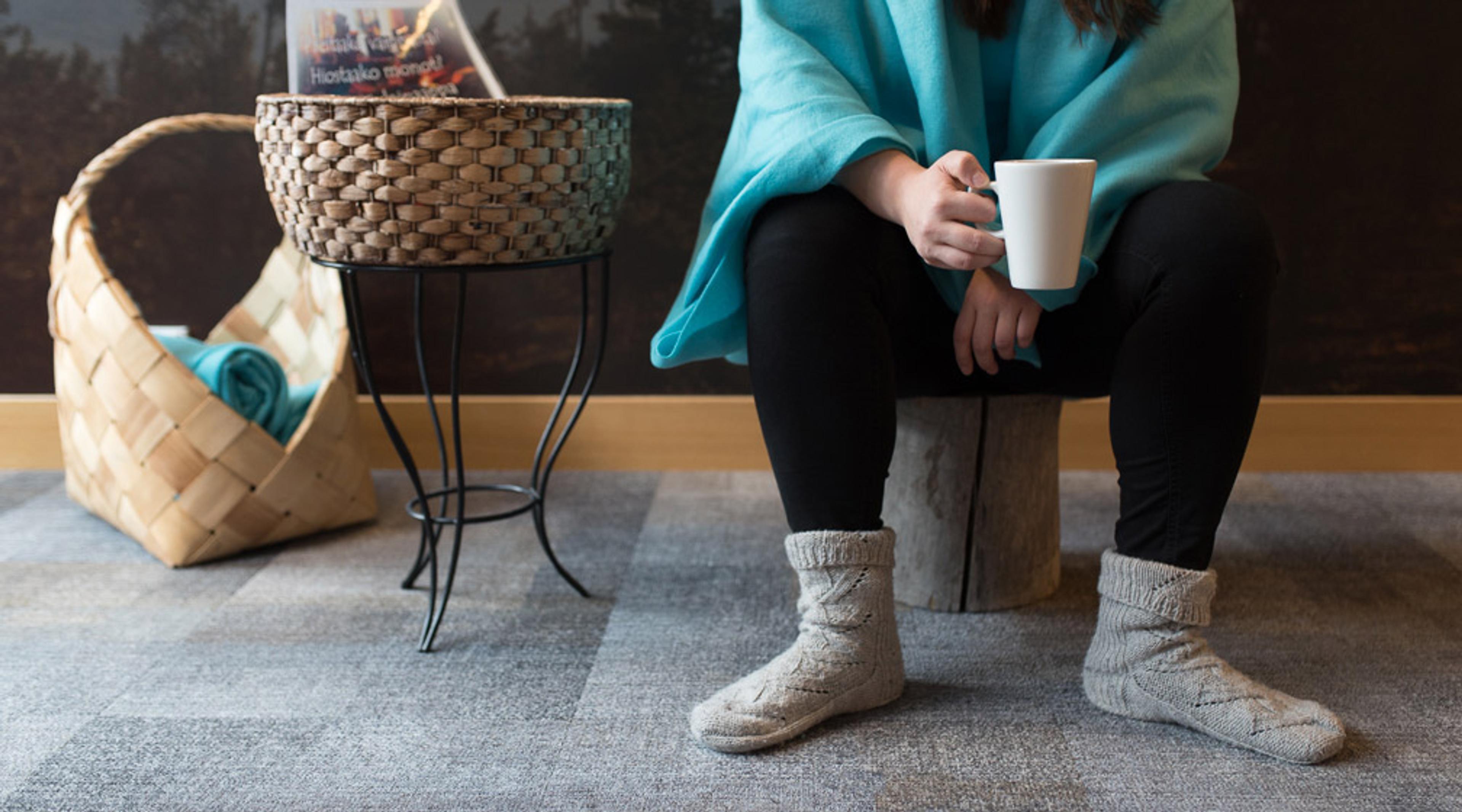 Person in turquoise blanket sitting on wooden stump holding white mug. Nearby wicker table and basket with blue towel.