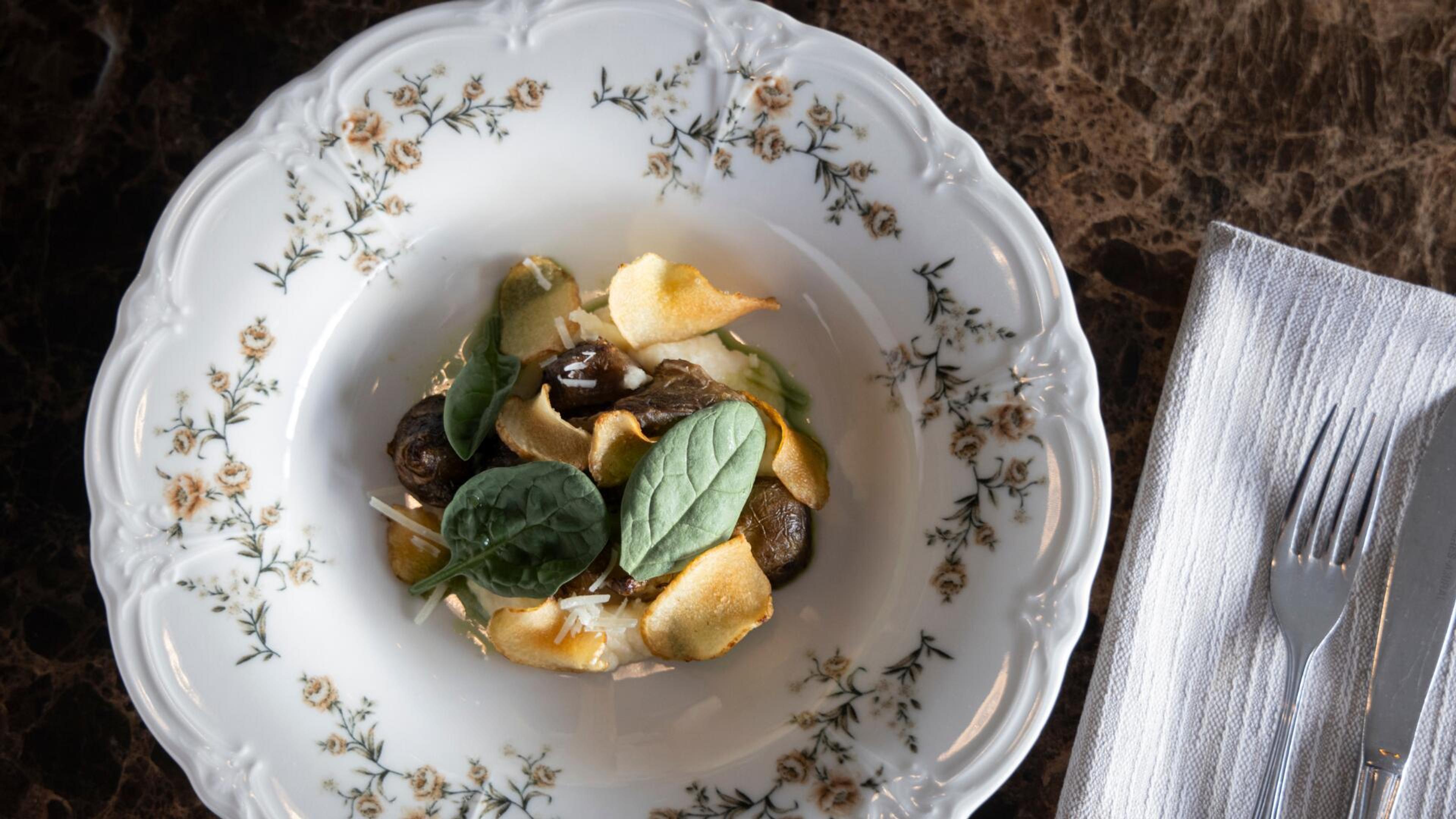 Koristeellinen posliinilautanen, jossa on annos sieniateriaa, pinaatinlehtiä ja perunalastuja. Vieressä valkoinen lautasliina ja haarukka.