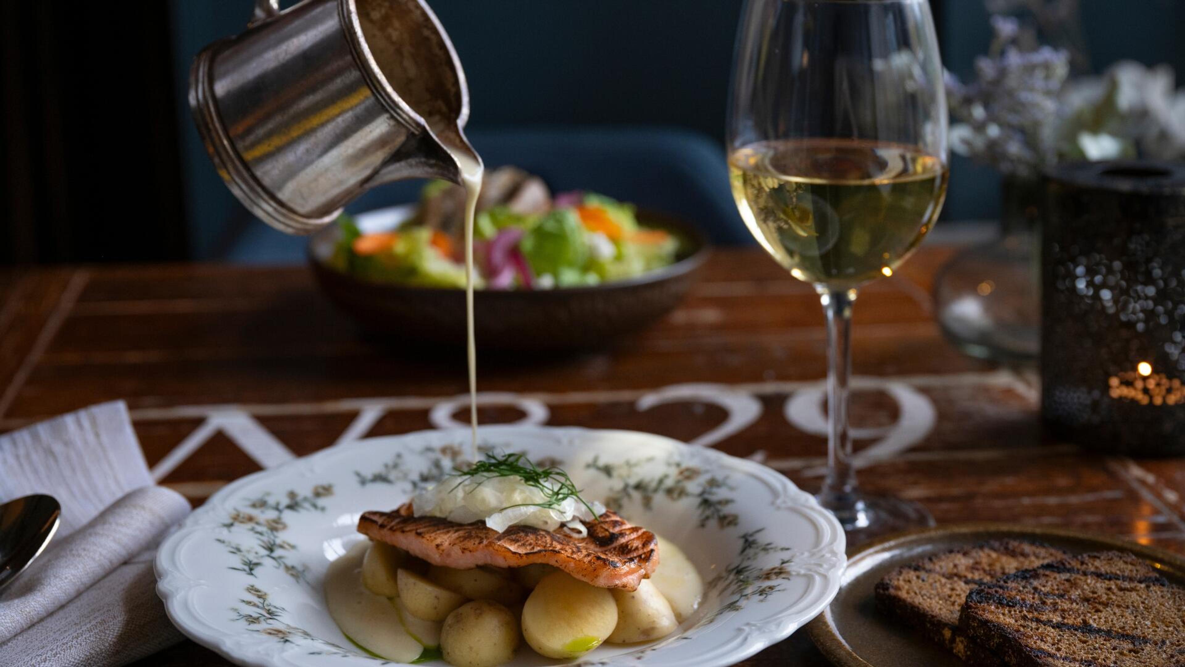 Metallikannusta kaadetaan kastiketta kala-annokselle, jossa on perunoita. Vieressä valkoviinilasi ja salaattia taustalla.