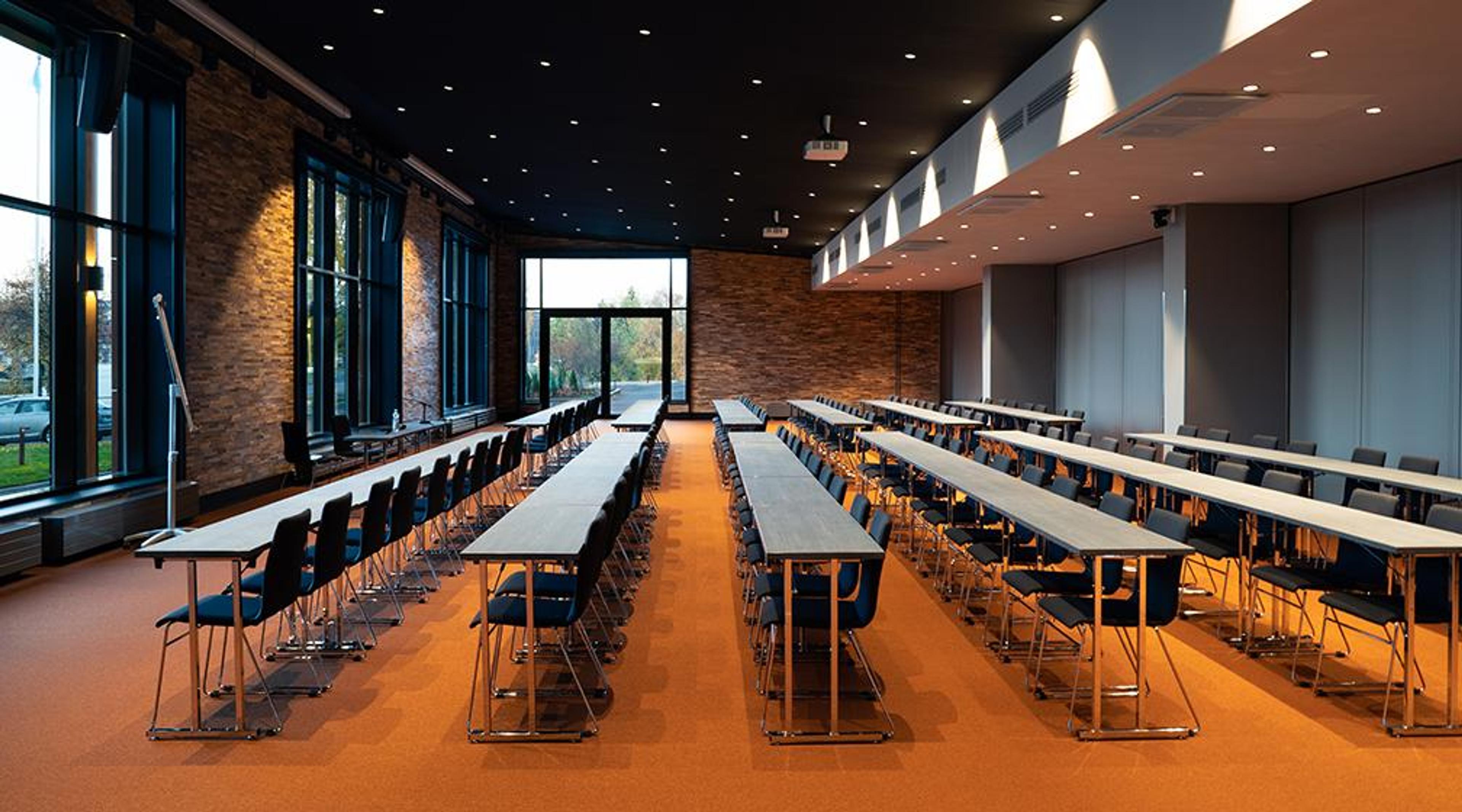 Spacious classroom with long tables, brick walls and large windows. Room has orange carpet flooring and blue chairs.