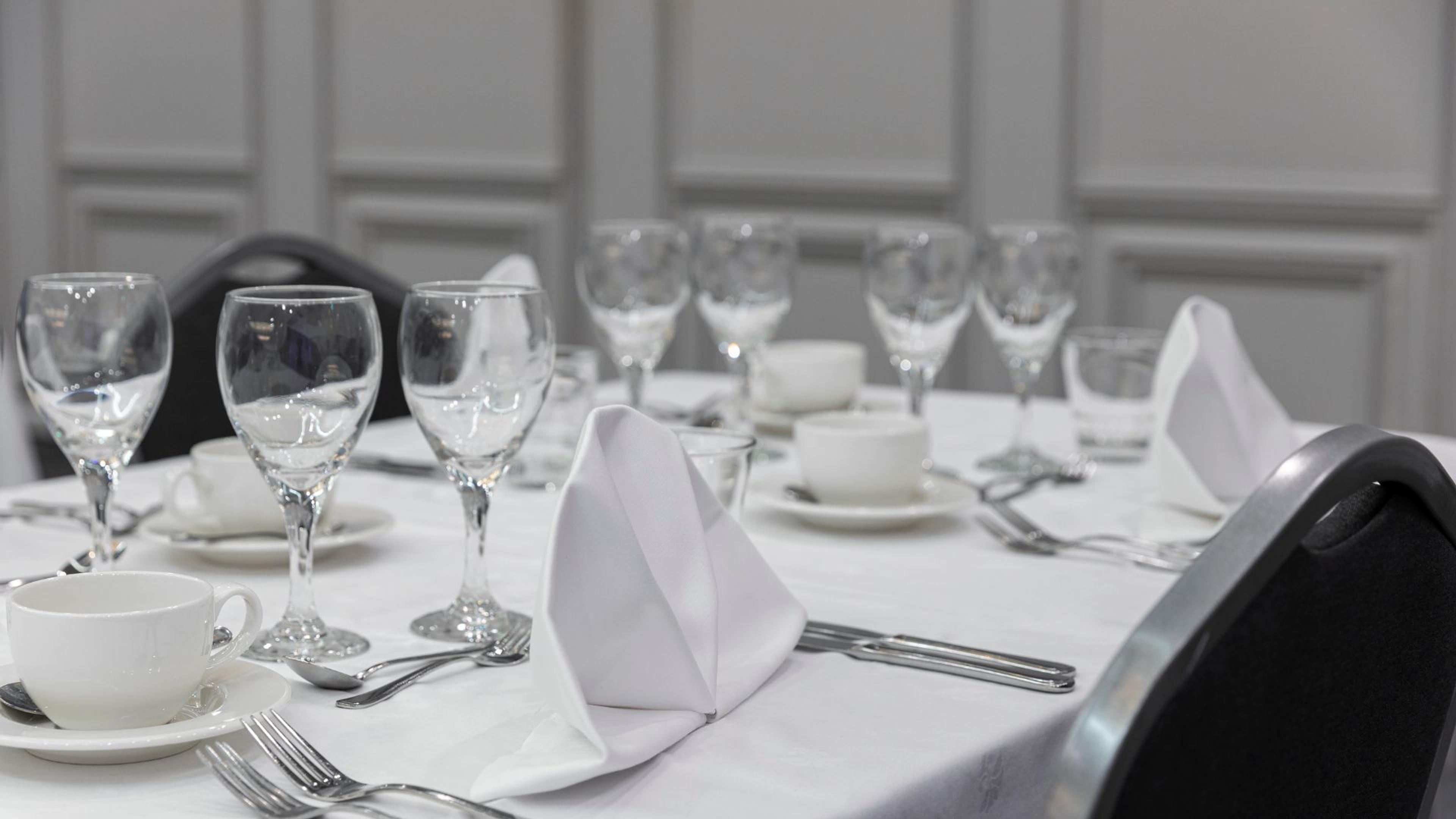 Elegantisti katettu pöytä valkoisella pöytäliinalla, viinilaseja, taitettu lautasliina, kahvikuppi ja ruokailuvälineet.
