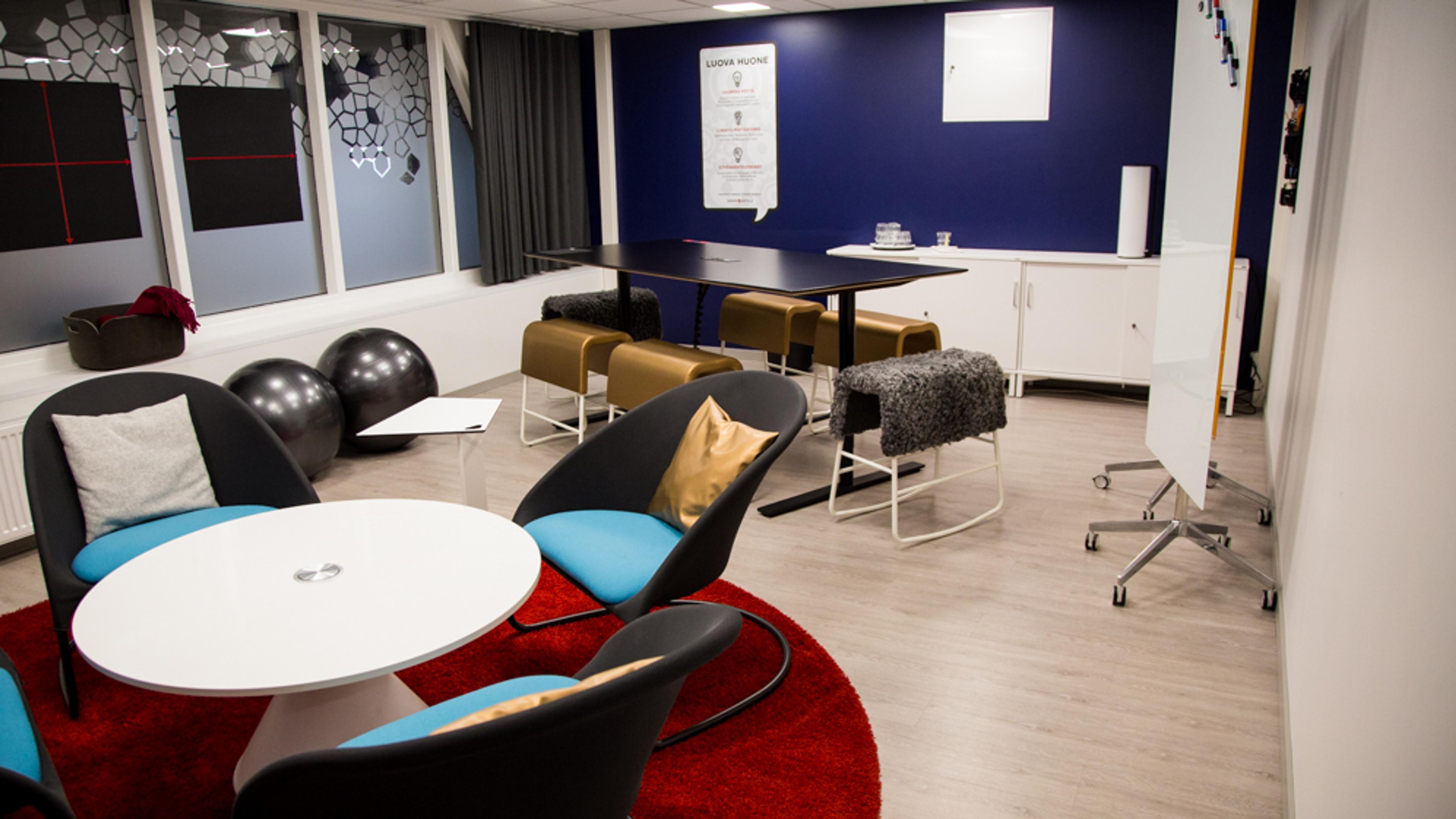 Modern meeting room with light flooring. White table with blue chairs on red rug and black table with brown chairs. Also exercise balls and a desk.
