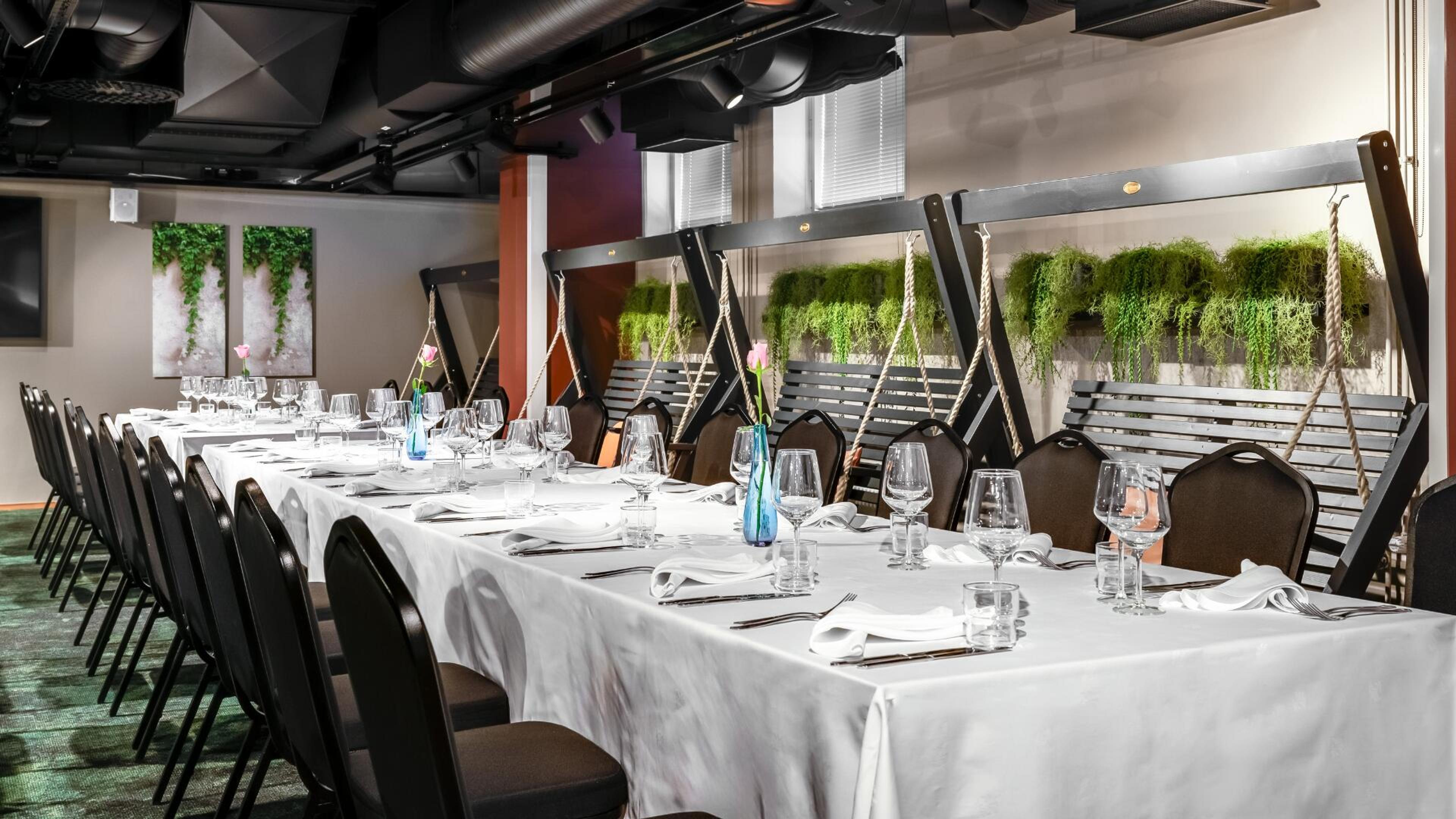 Moderni ravintolasali, jossa pitkä pöytä valkoisella liinalla ja koristeelliset keinut seinän vieressä. Tila on koristeltu roikkuvilla viherkasveilla.