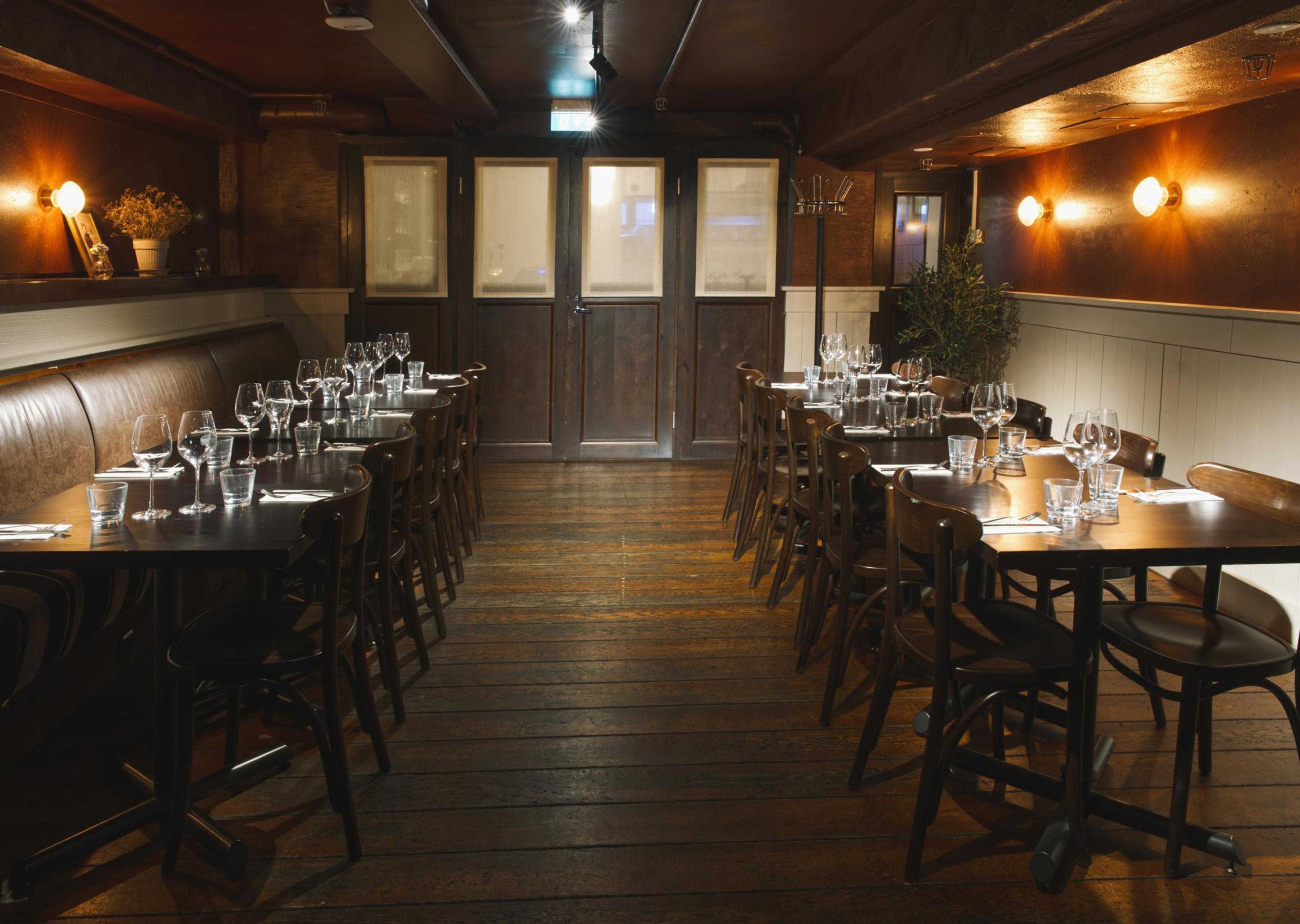 Tunnelmallinen ravintolan kabinetti puisella lattialla, jossa pöydät katettu lasein ja aterimiin. Lämmin valaistus seinävalaisimista.
