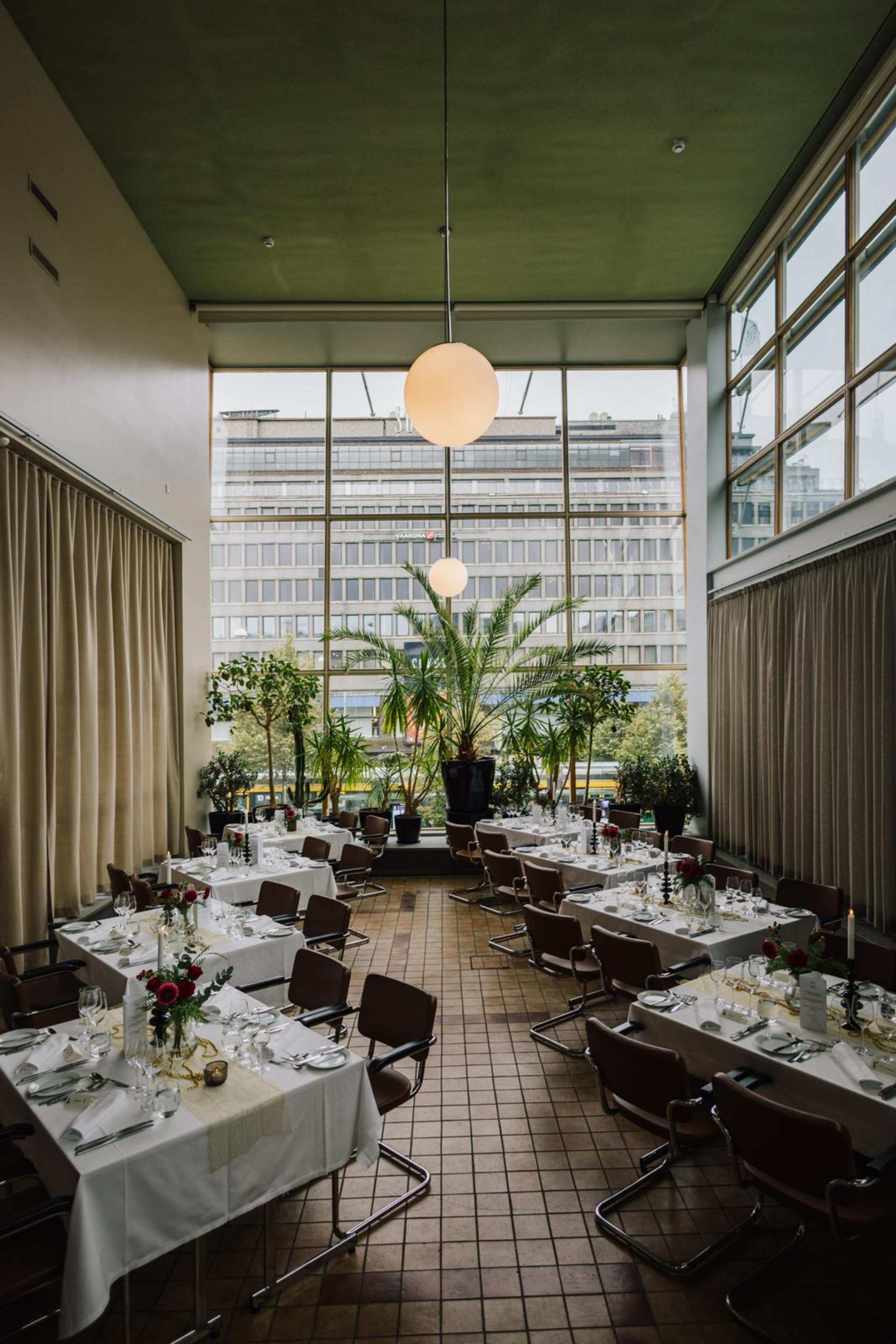 Valoisa ravintola korkeilla ikkunoilla. 8 henkilön pöytäryhmiä valkoisilla liinoilla, taustalla viherkasveja. Laattalattia ja pyöreät riippuvalaisimet.