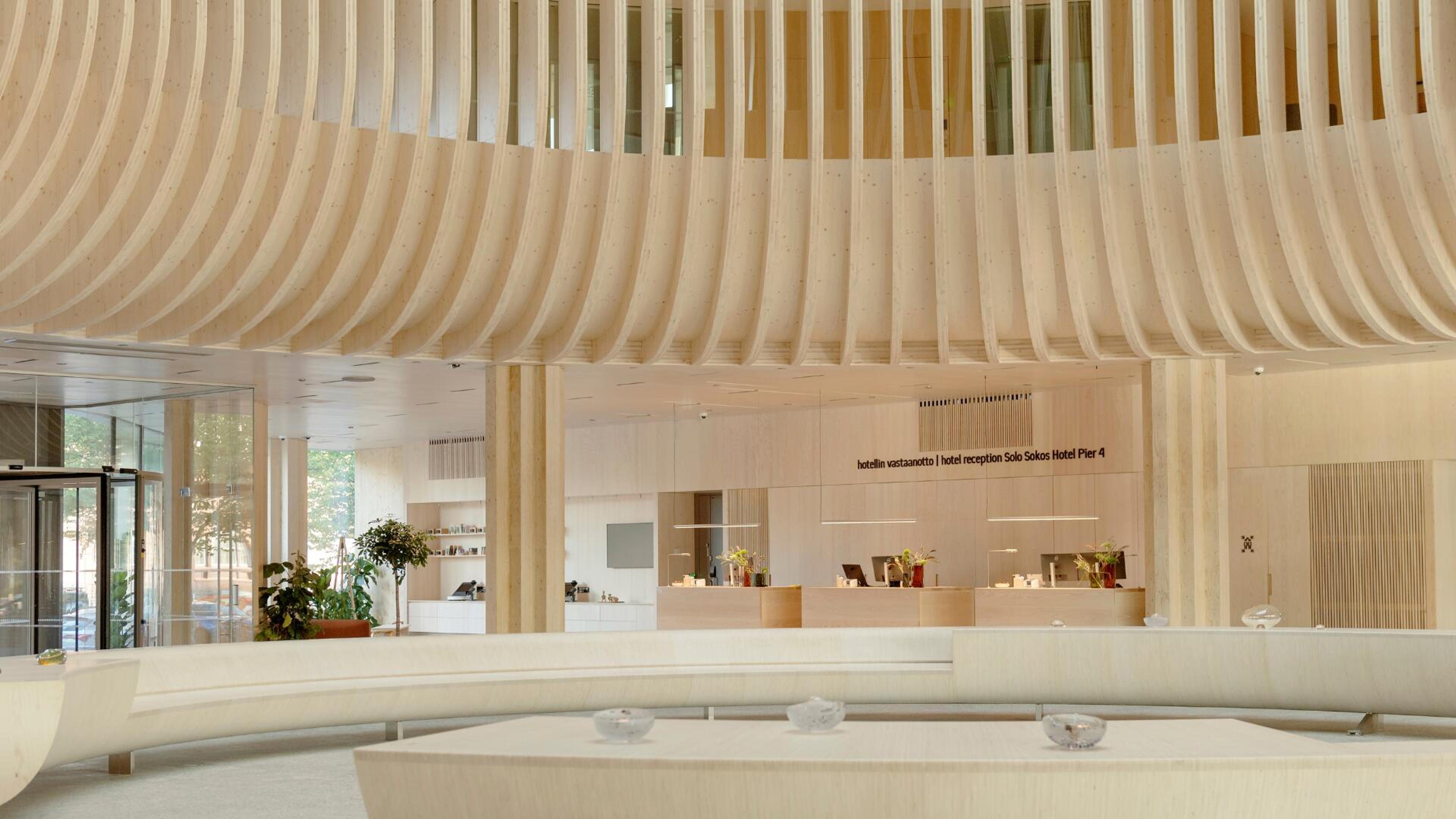 Moderni hotellin aula, jossa vaalea puinen kaareva katto, vastaanottotiski ja lasiseinät. Avara tila luonnonvalolla ja viherkasveilla.