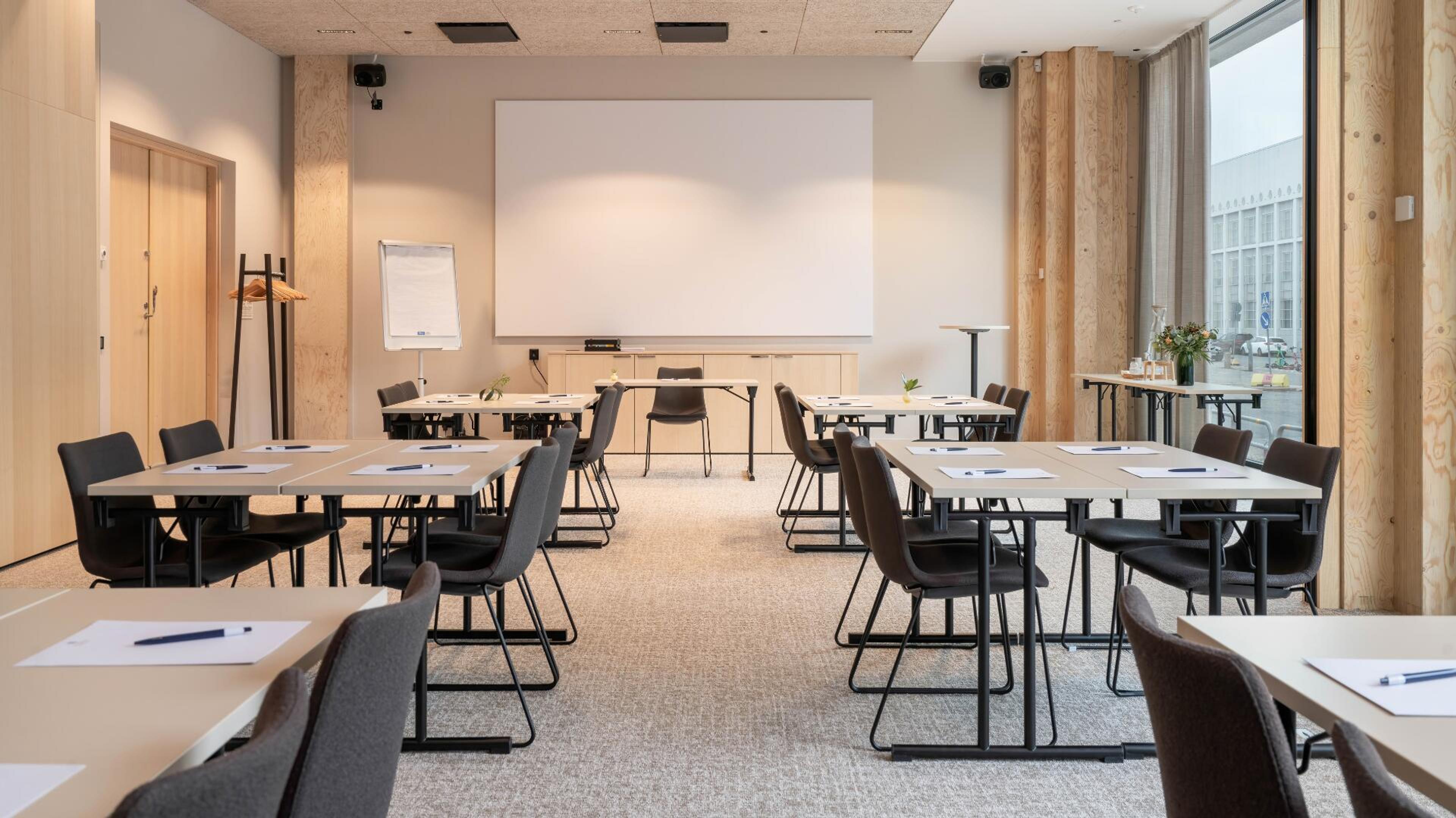 Moderni kokoushuone vaalean puisilla pinnoilla, jossa pöydät ja tuolit ryhmitelty luokkahuonemaisesti. Tilassa projektorikangas ja lattialla lyhytnukkainen kokolattiamatto.