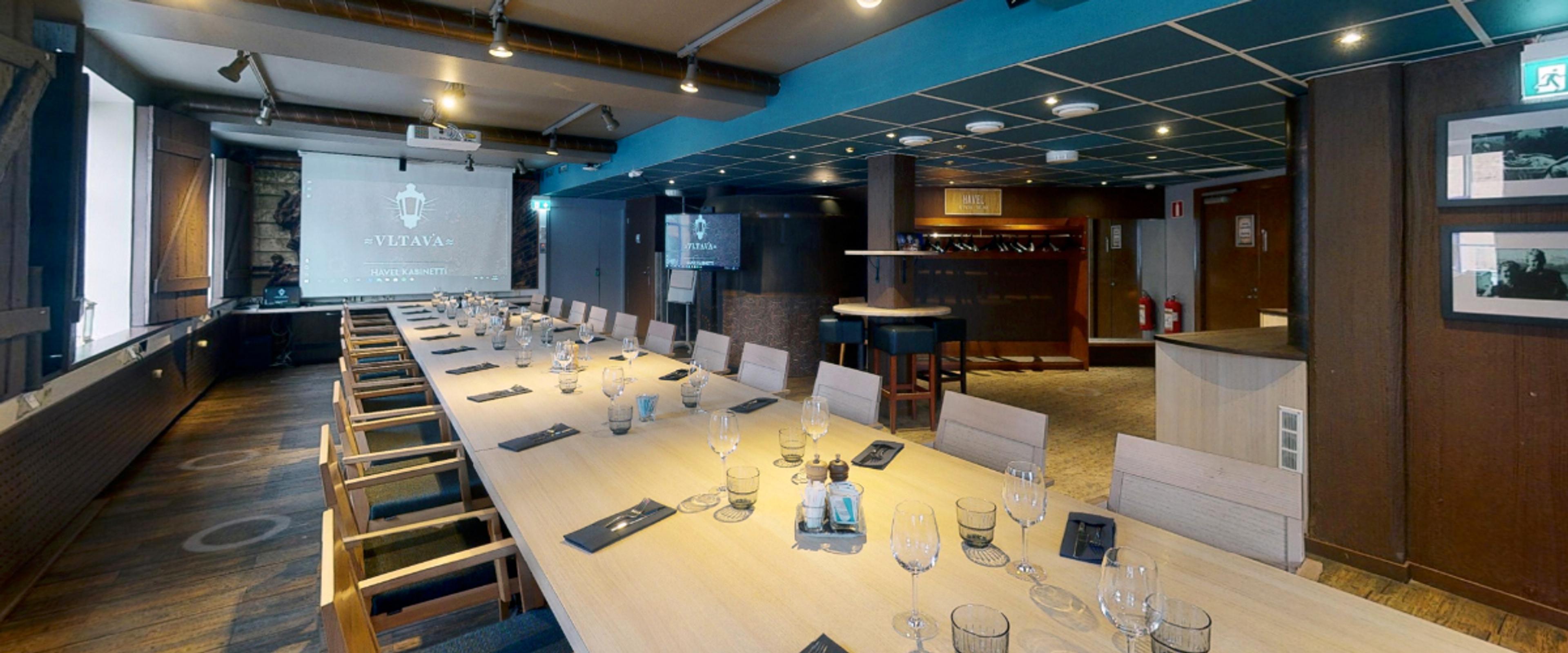 Moderni kokoustila pitkällä puisella pöydällä, jonka ympärillä on tuoleja. Taustalla on valkokangas, naulakko ja pieni keittiö.