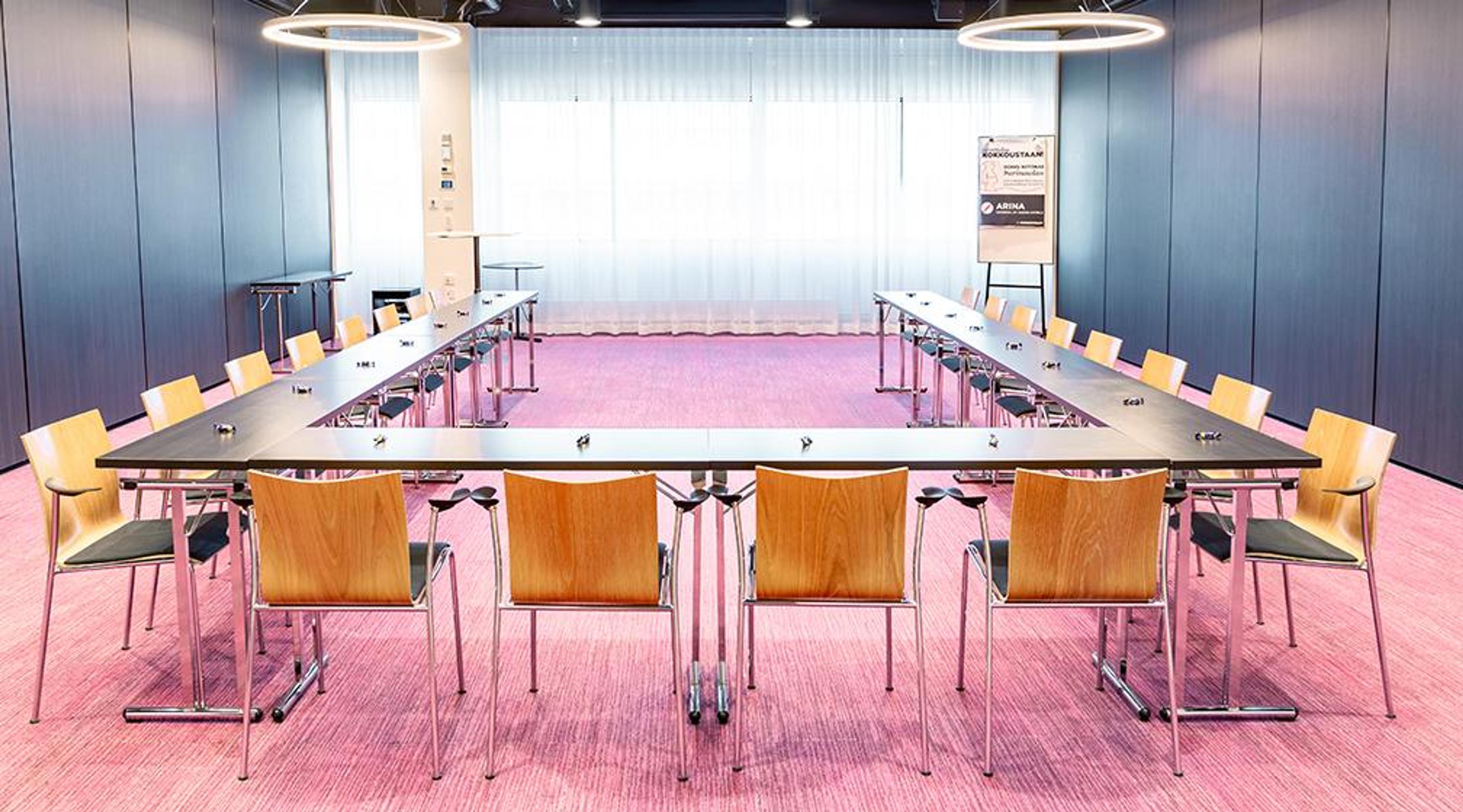 Kokoushuone U-muotoisella pöytäjärjestelyllä, puiset tuolit, vaaleanpunainen kokolattiamatto ja modernit rengasvalaisimet katossa.
